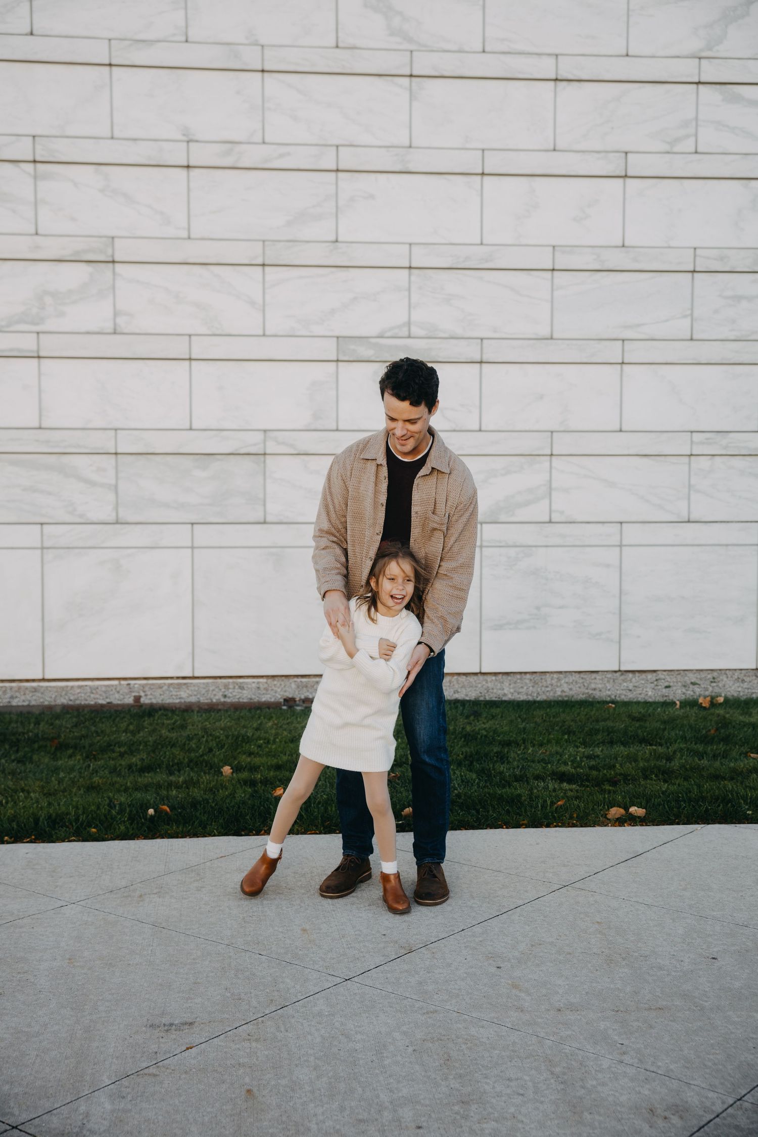 A father and young daughter smile and play together outside a white marble building with green grass.