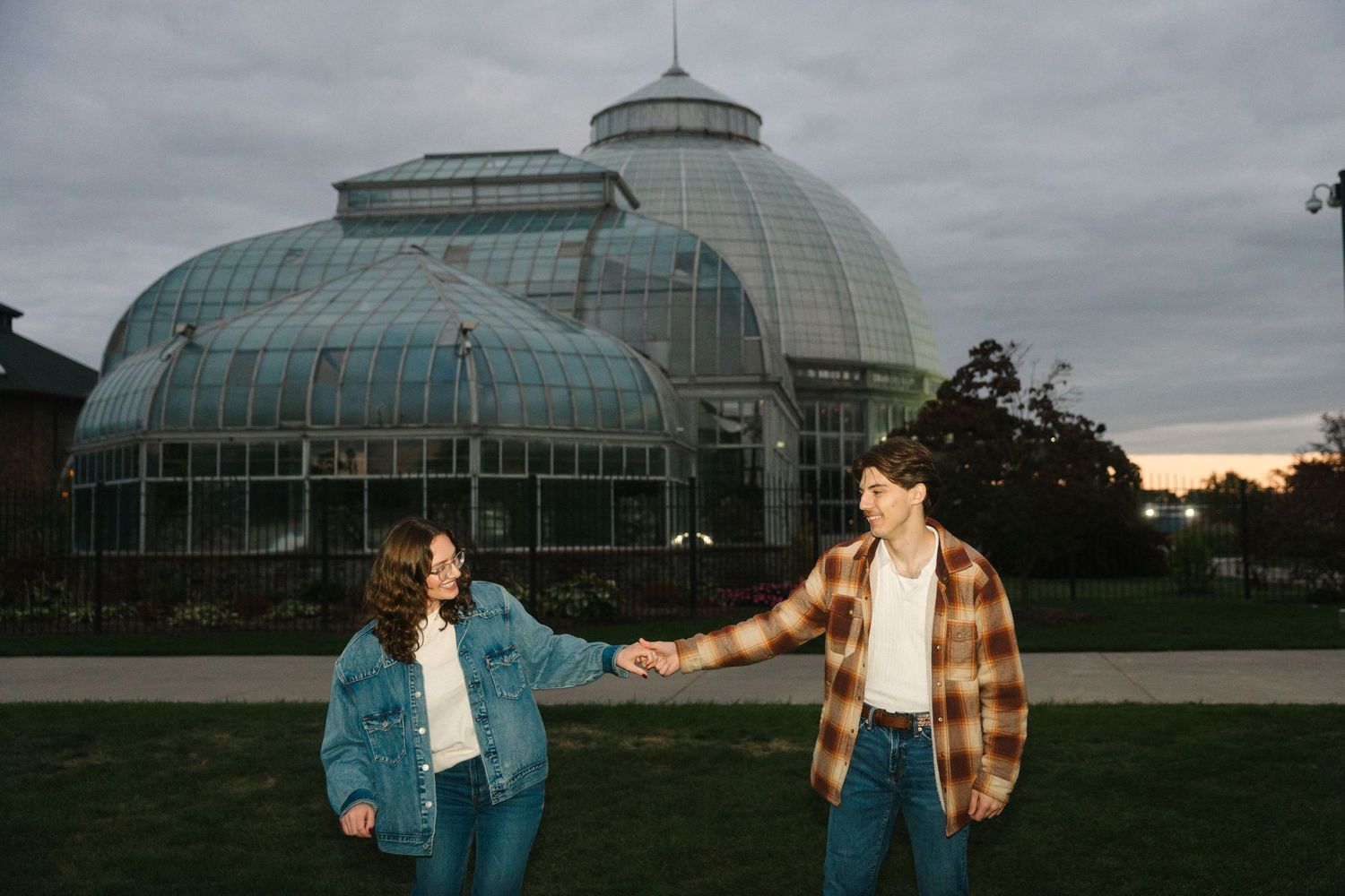 Couple holding hands in front of a large glass greenhouse conservatory at dusk, wearing denim and plaid.
