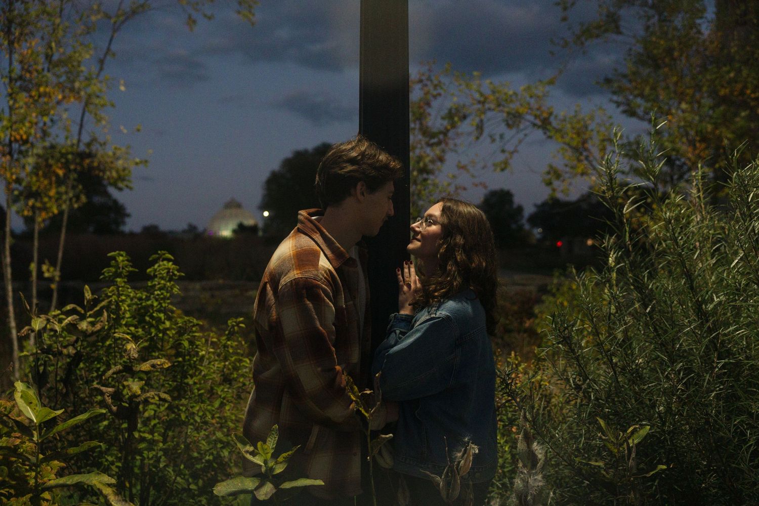 A couple shares a romantic moment surrounded by lush greenery at dusk in a cinematic outdoor scene.