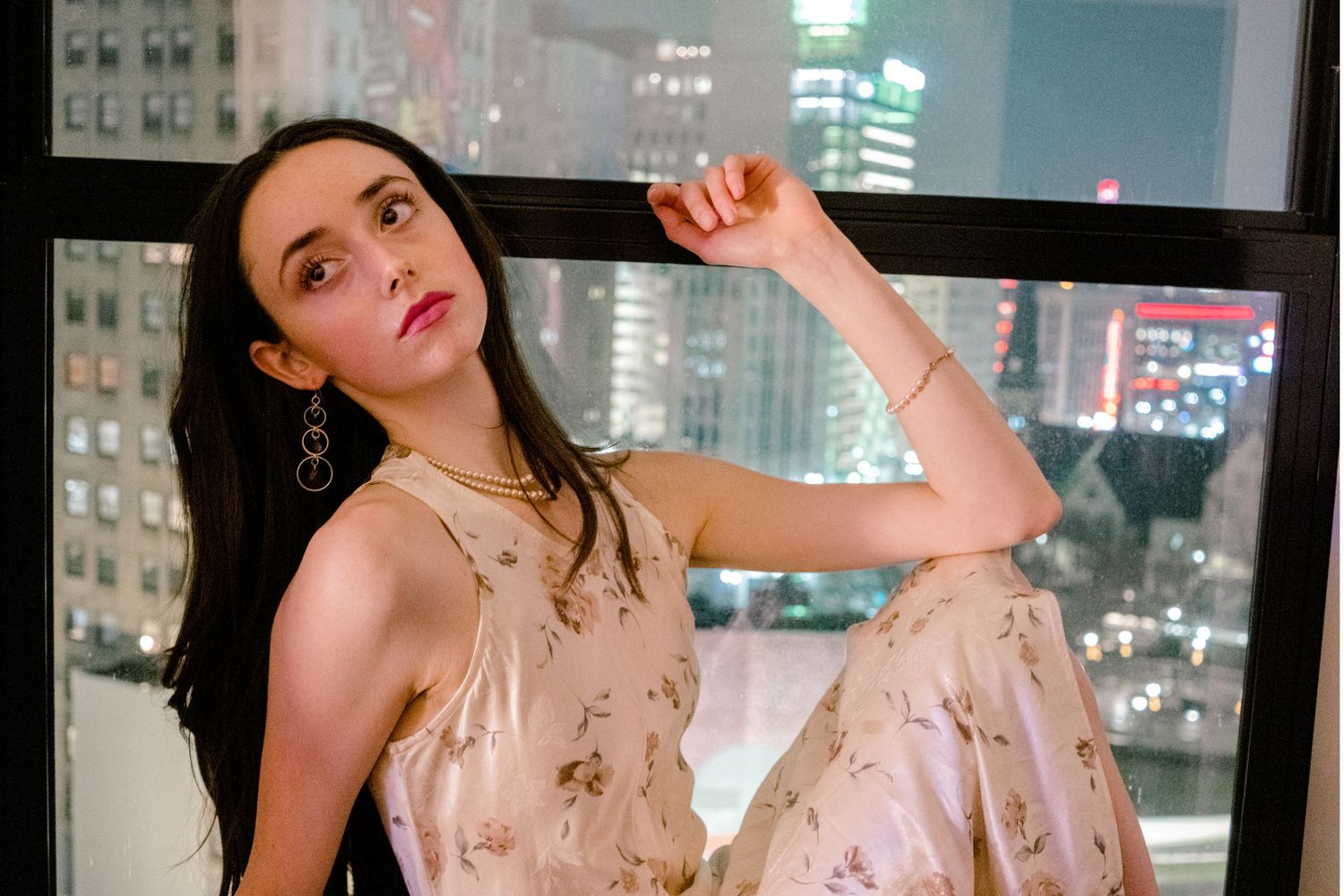 Young woman in floral dress poses elegantly by city window at night with glowing skyline backdrop.