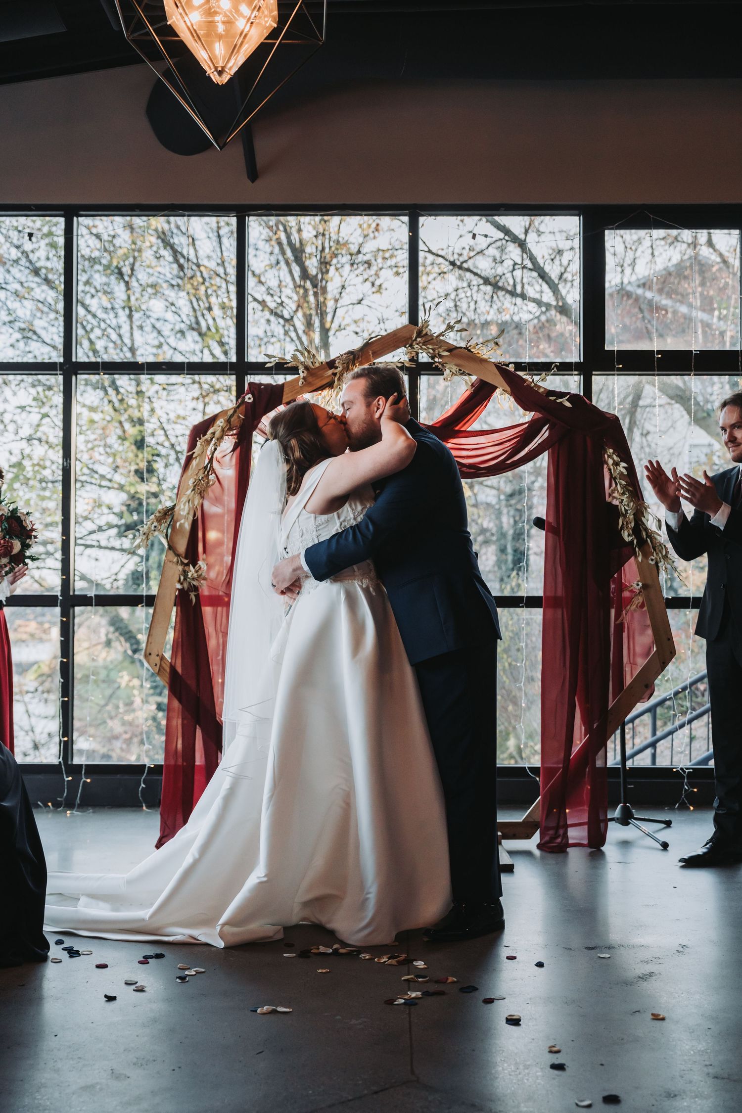 Bride and groom share first kiss under burgundy draped arch during indoor wedding ceremony.