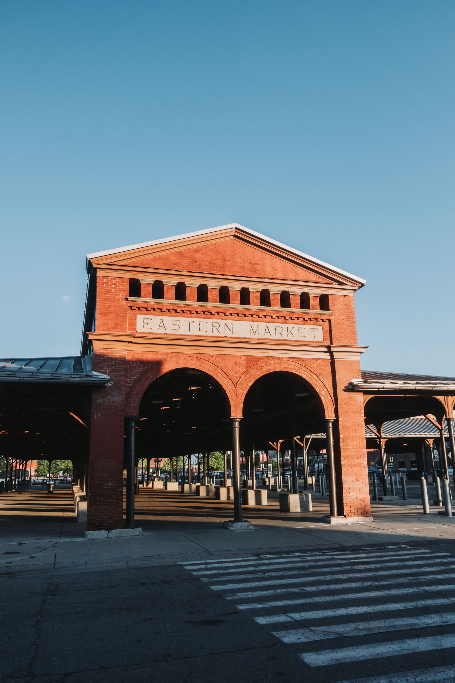 Red brick Eastern Market building with arched entrance and covered outdoor stalls under clear blue sky in Washington DC.