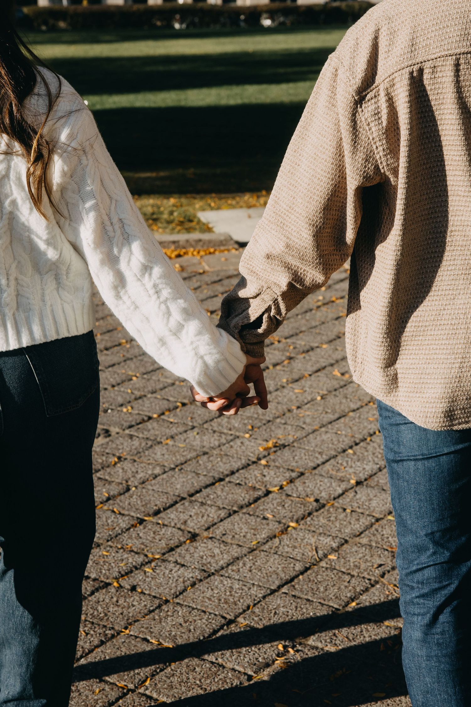 Couple holding hands while walking on a brick path in autumn, wearing casual sweaters and jeans.