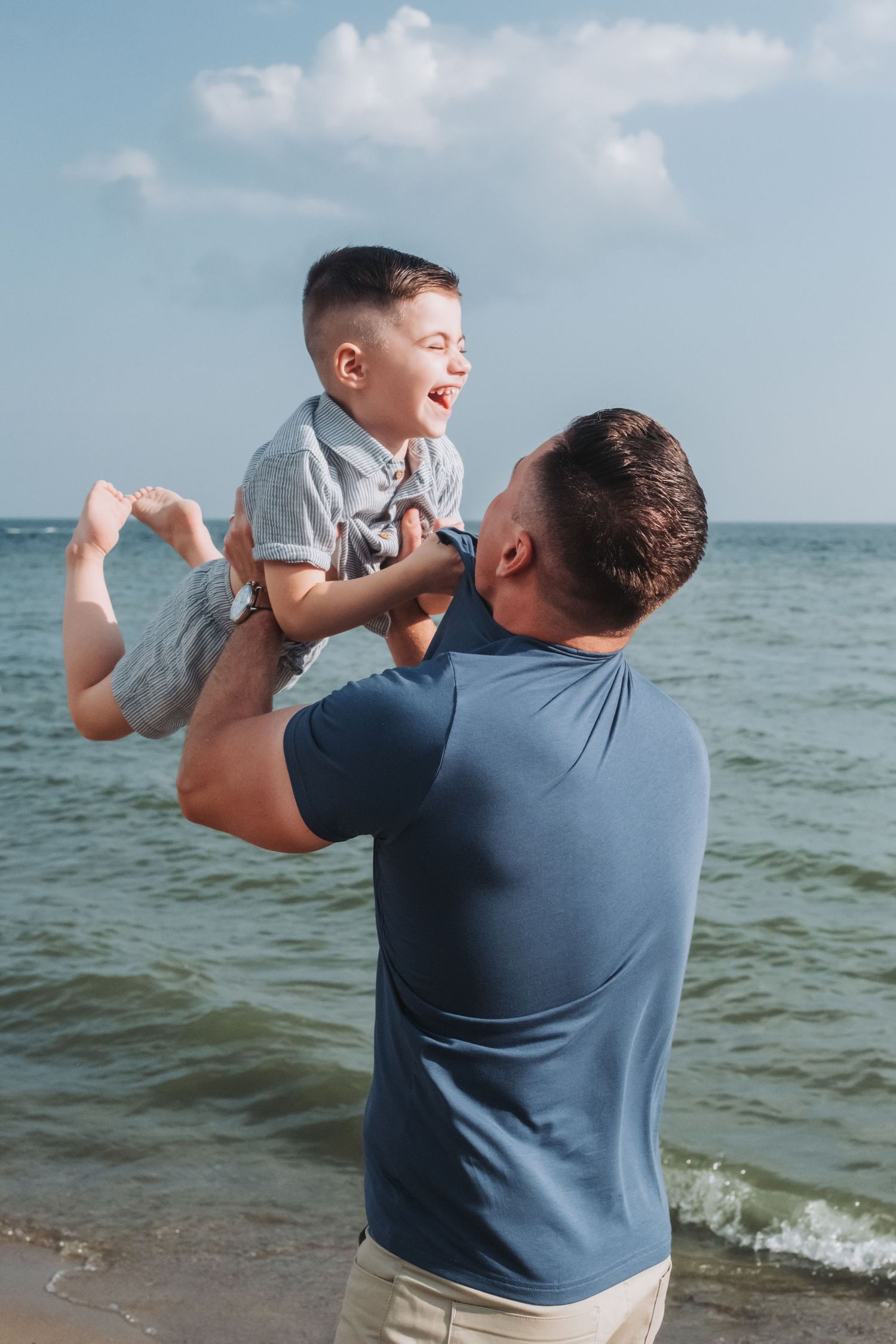 Father joyfully lifts laughing son at the beach with ocean waves and blue sky in the background.
