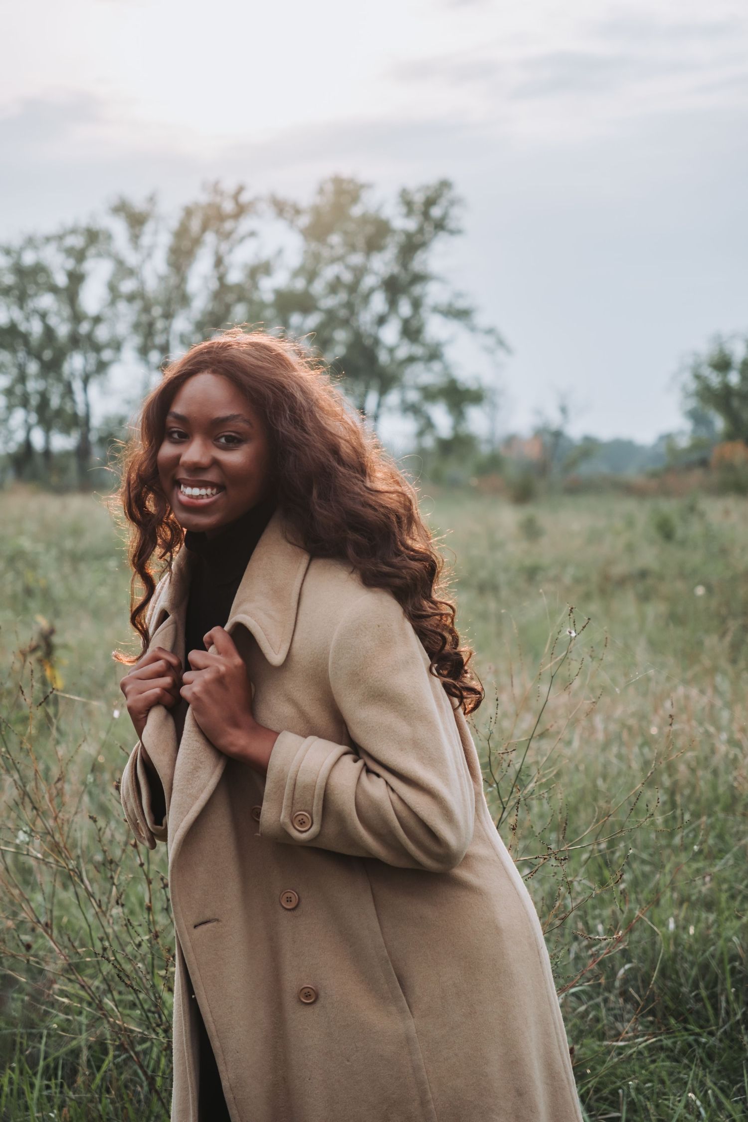Smiling Black woman with curly auburn hair wearing a beige trench coat standing in a grassy field.