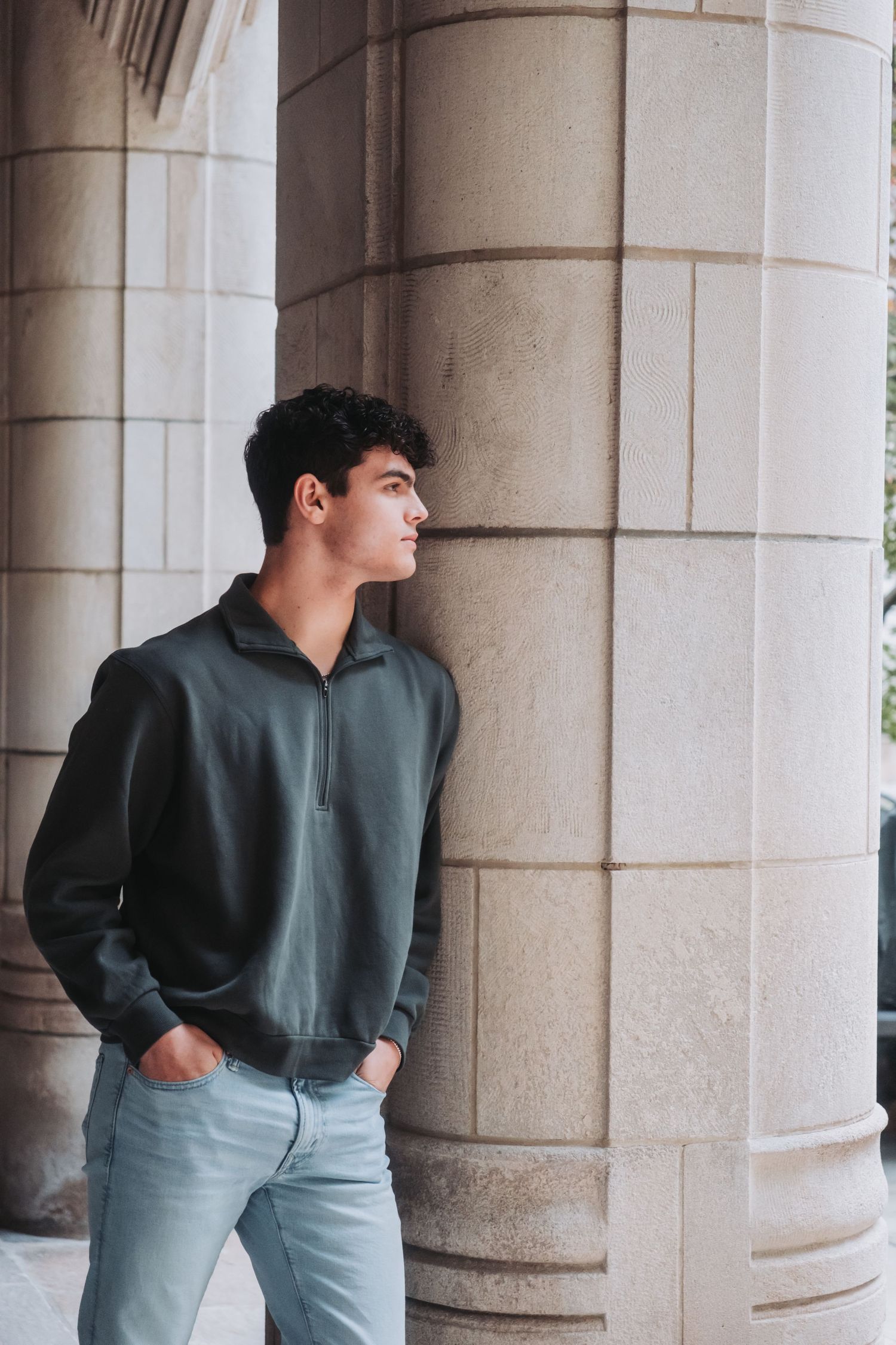 Person in dark sweater poses against stone columns in architectural setting.