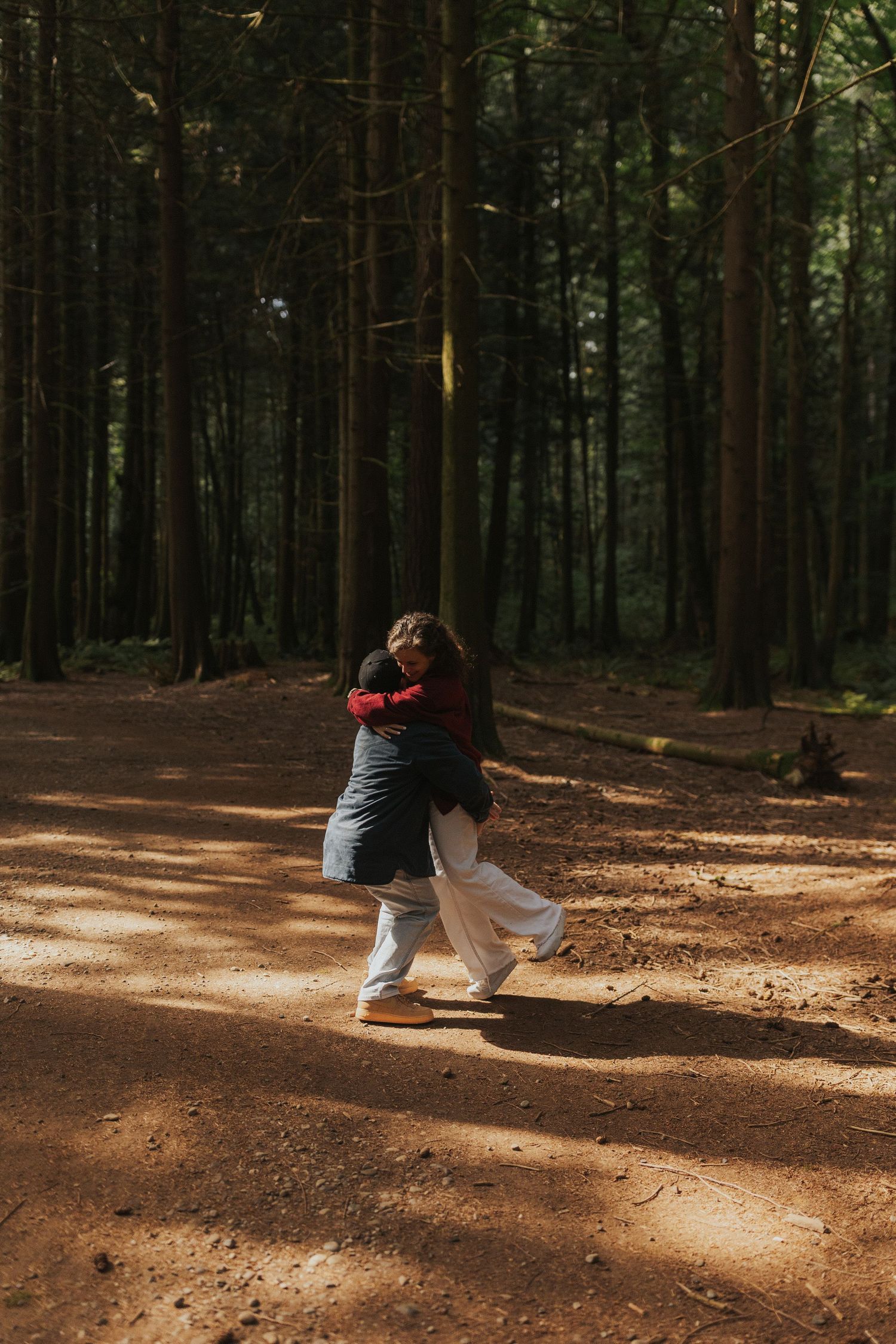Dancing figures cast shadows on a sunlit forest floor surrounded by tall trees.