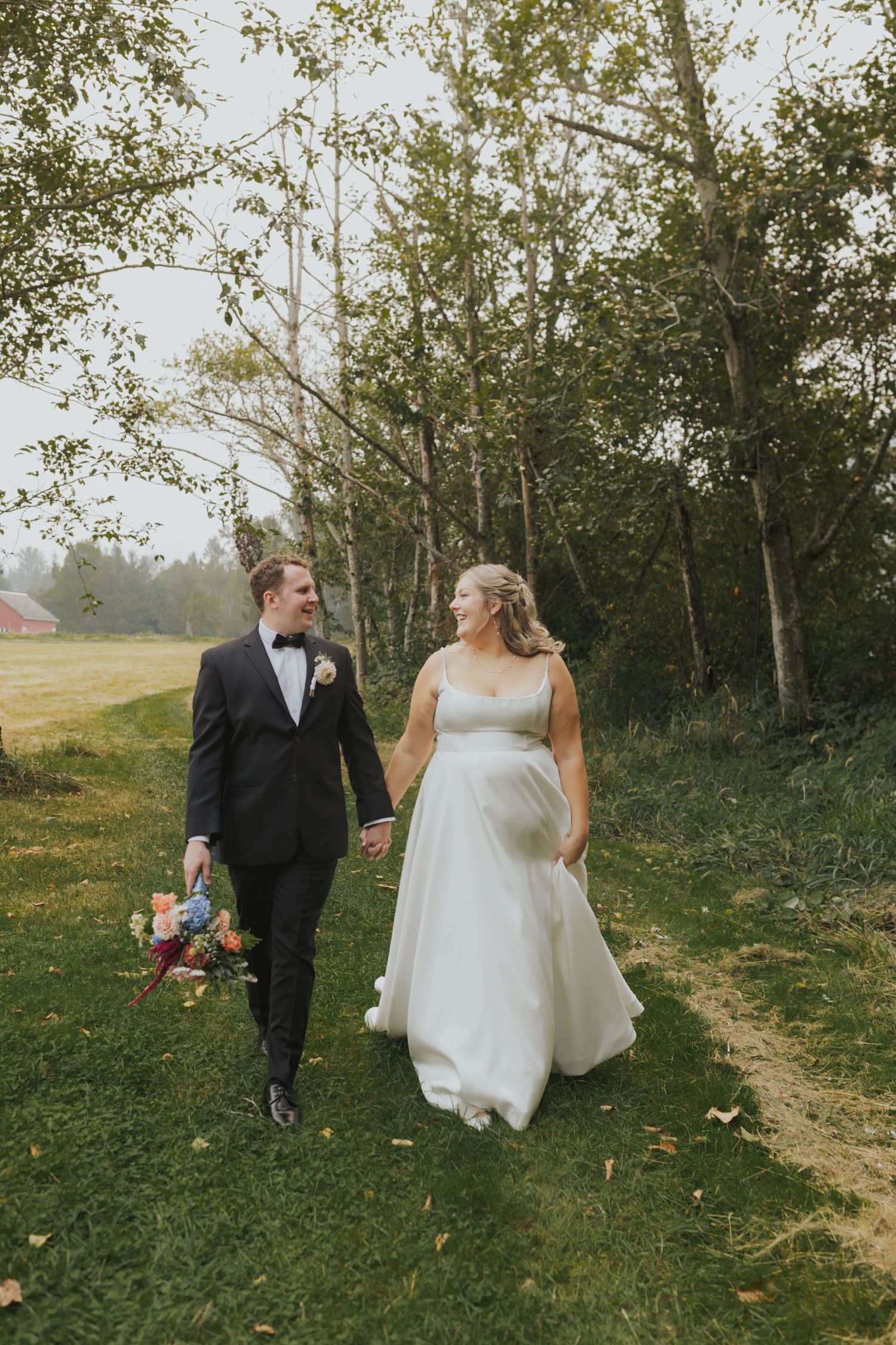 A newlywed couple walks hand in hand through a scenic outdoor setting with fall foliage.