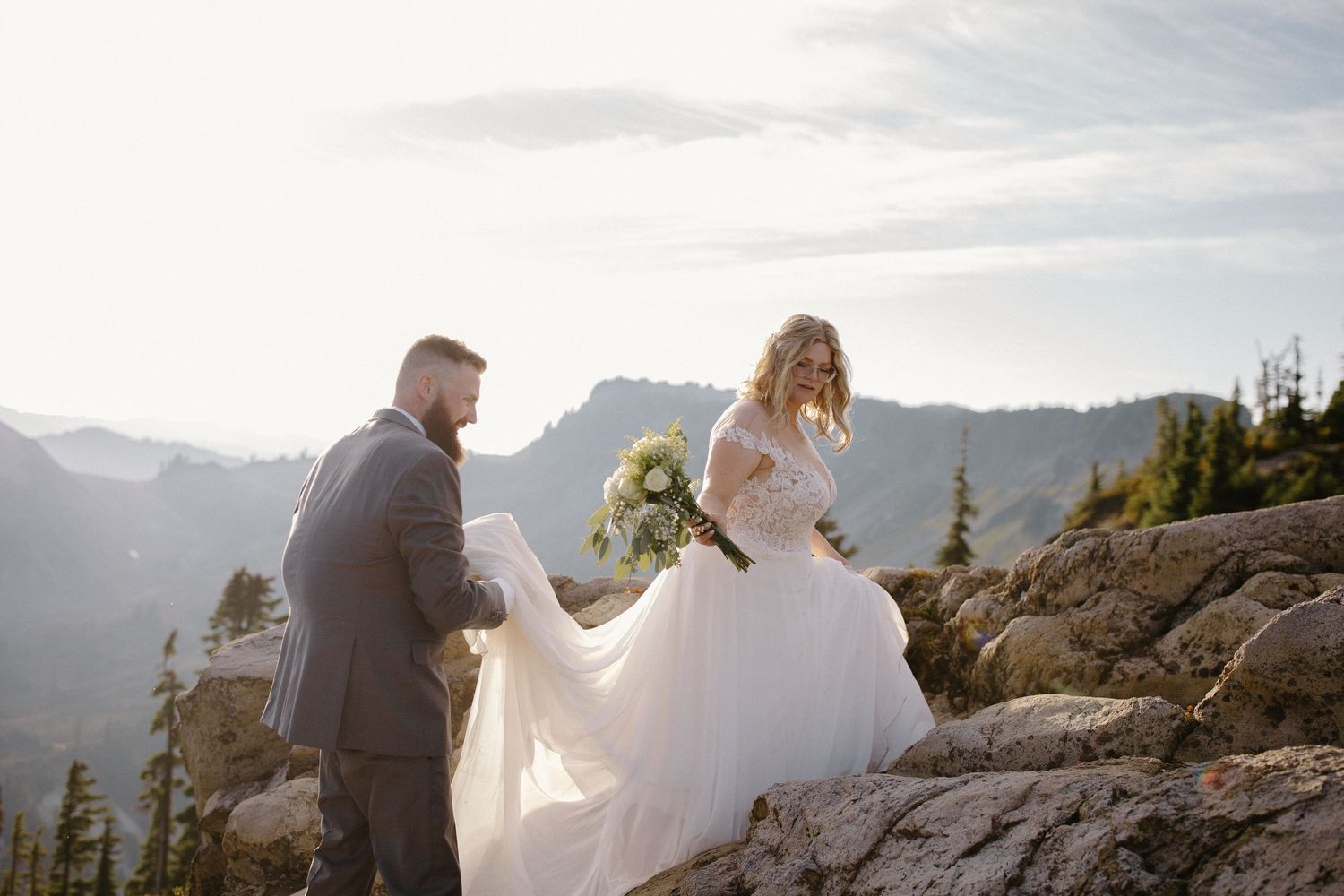 A couple in wedding attire poses dramatically on rocky mountain terrain at sunset.