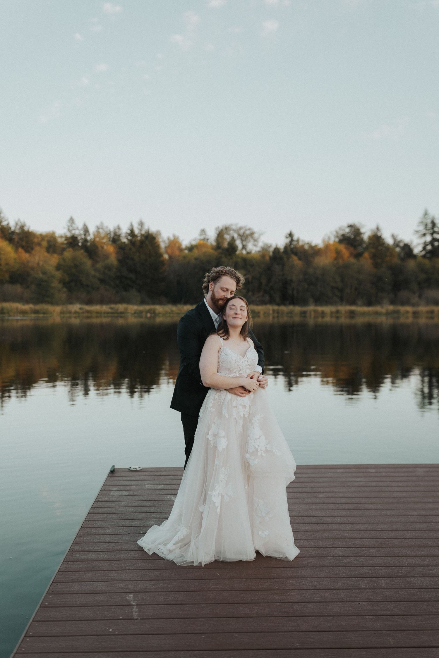 A romantic wedding couple sits together on a wooden dock overlooking a serene lake during golden hour.