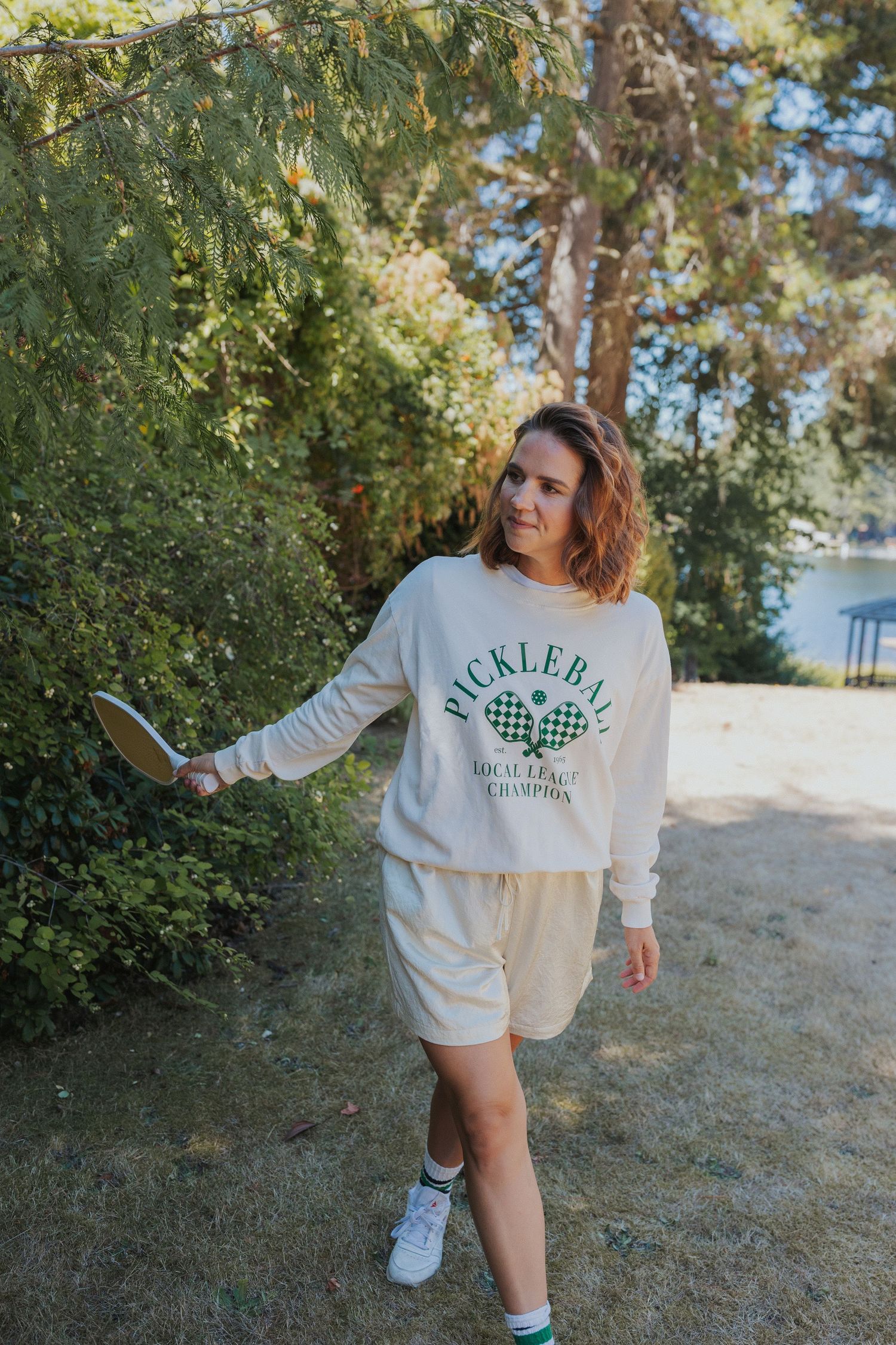 Person wearing an oversized white Pickleball Champion sweatshirt poses outdoors near trees.