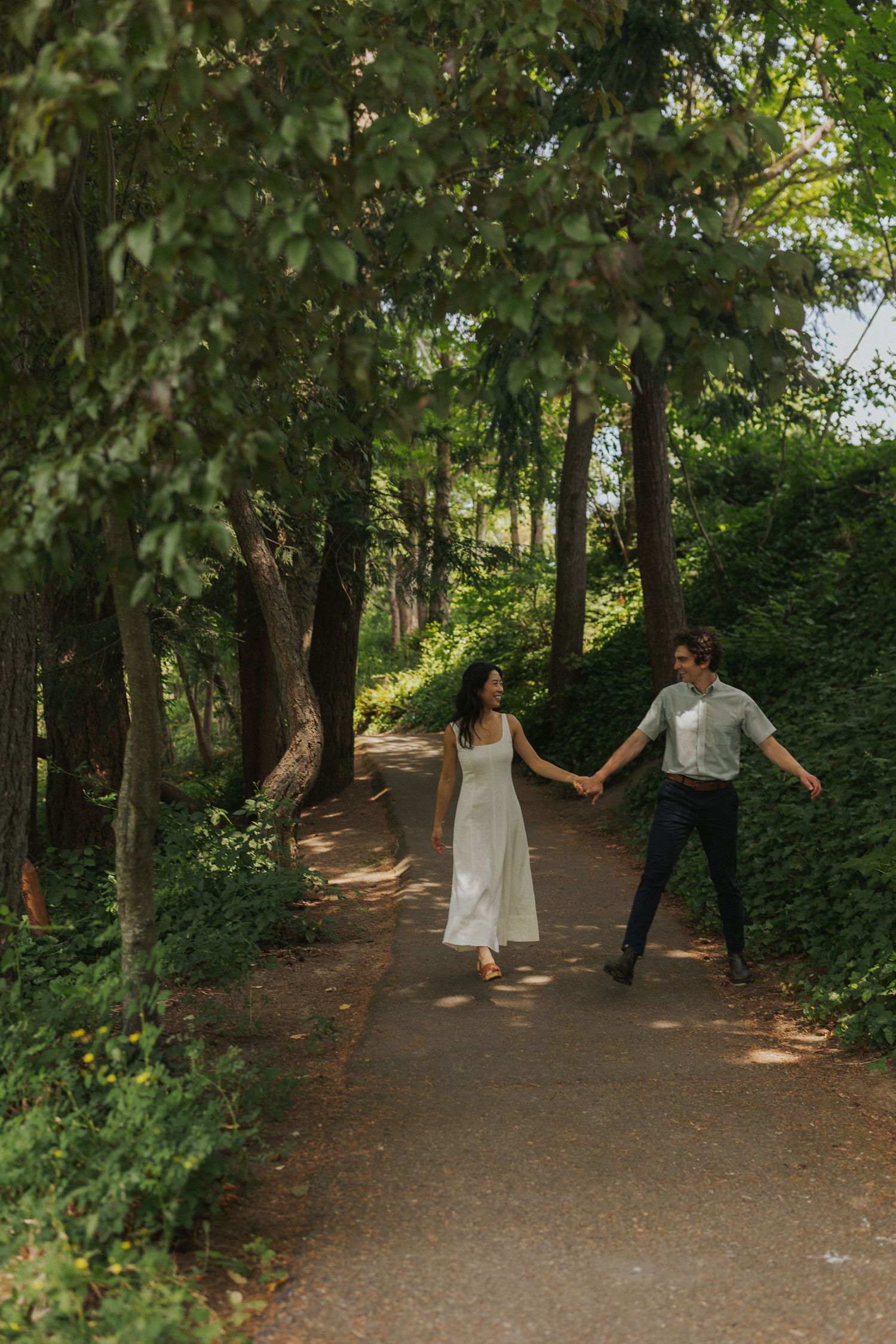 Couple walking and dancing together on a tree-lined path in a romantic outdoor photoshoot.