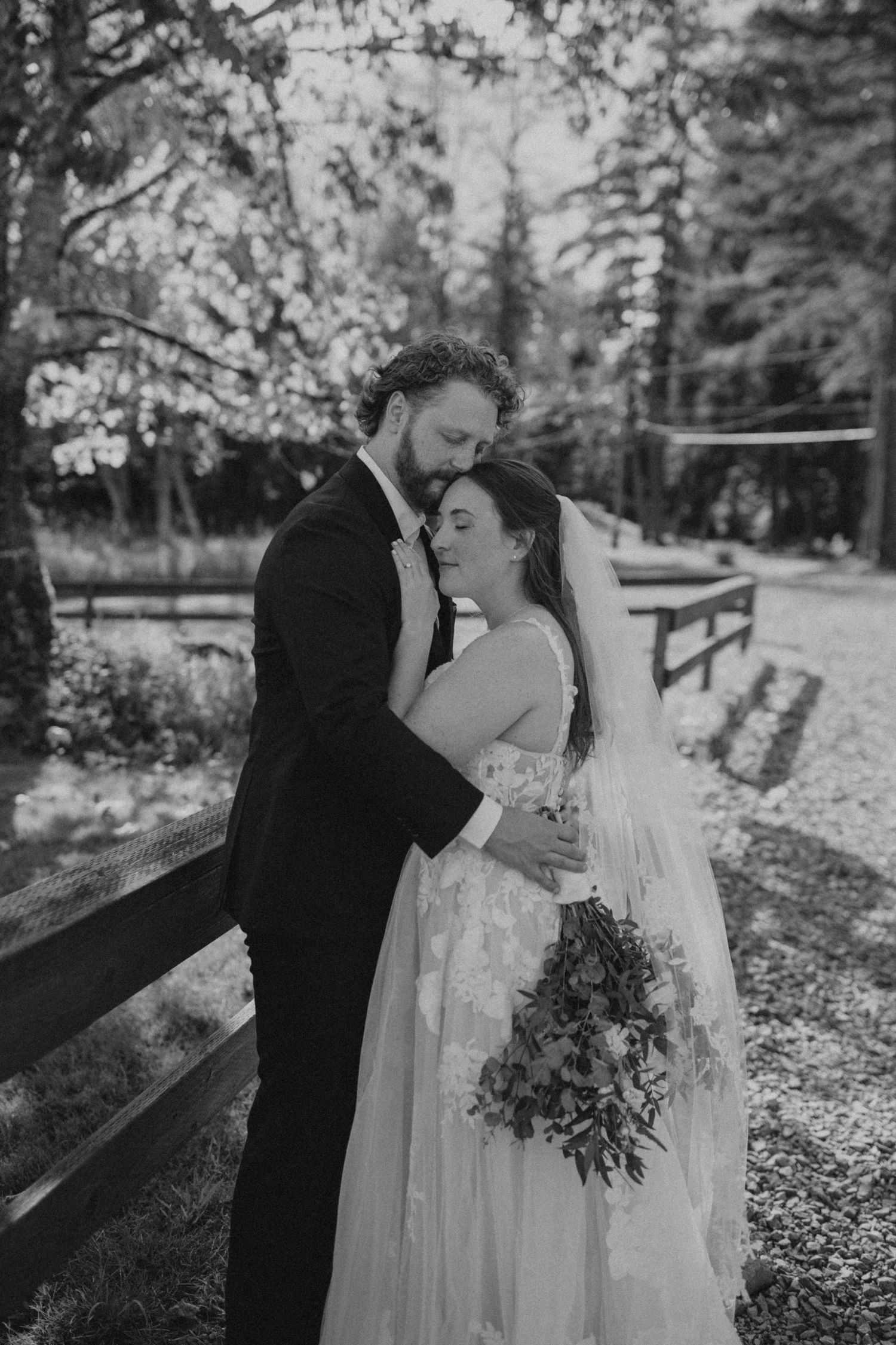 Black and white romantic wedding portraits taken along a rustic fence in a wooded setting.