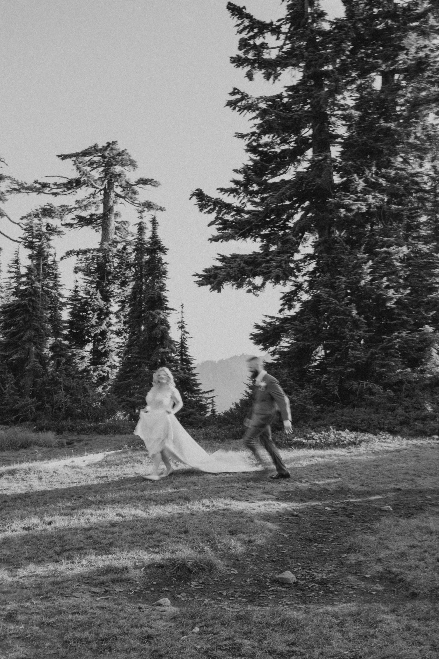 A black and white photo of a couple dancing joyfully among tall evergreen trees.