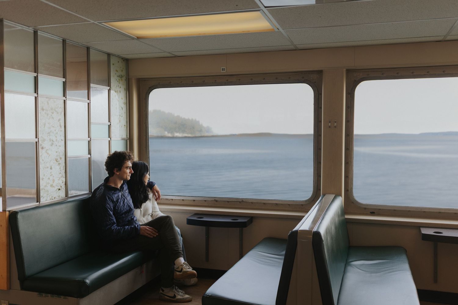 Interior view of ferry boat cabin with benches and large windows overlooking water.