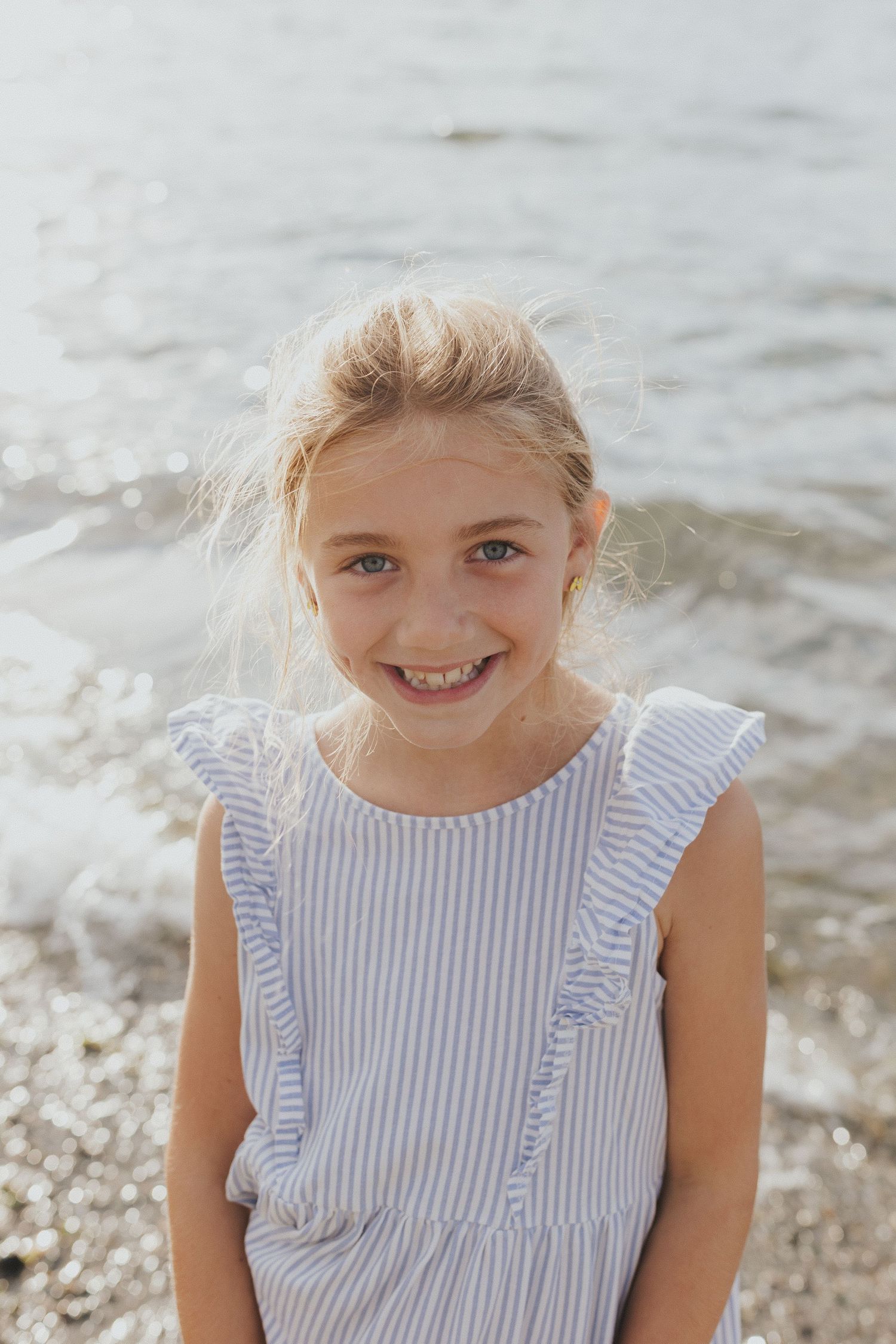 Young child in striped ruffled top smiling by the water's edge.
