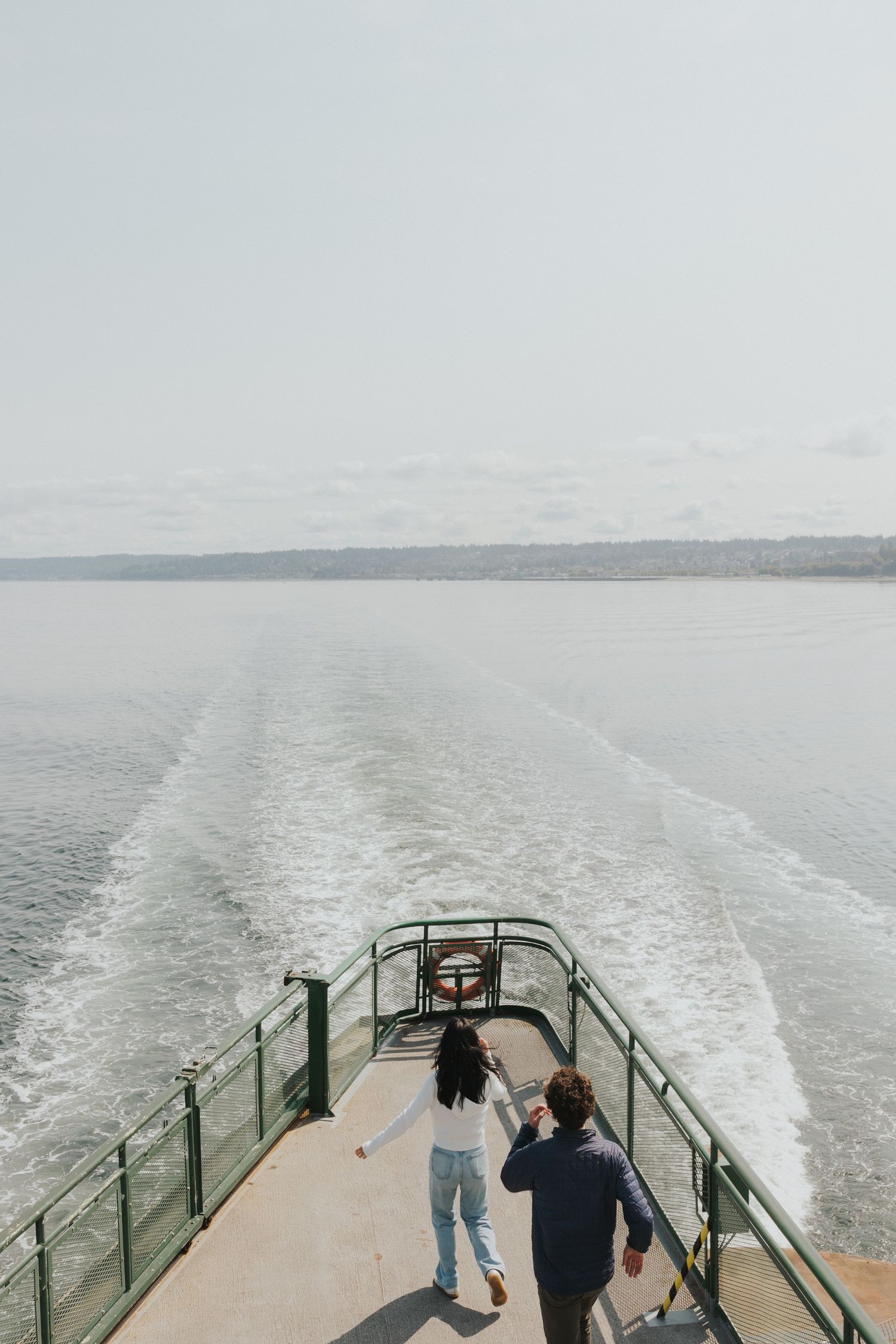 Two people stand at the back of a ferry boat watching the wake trail in the water.