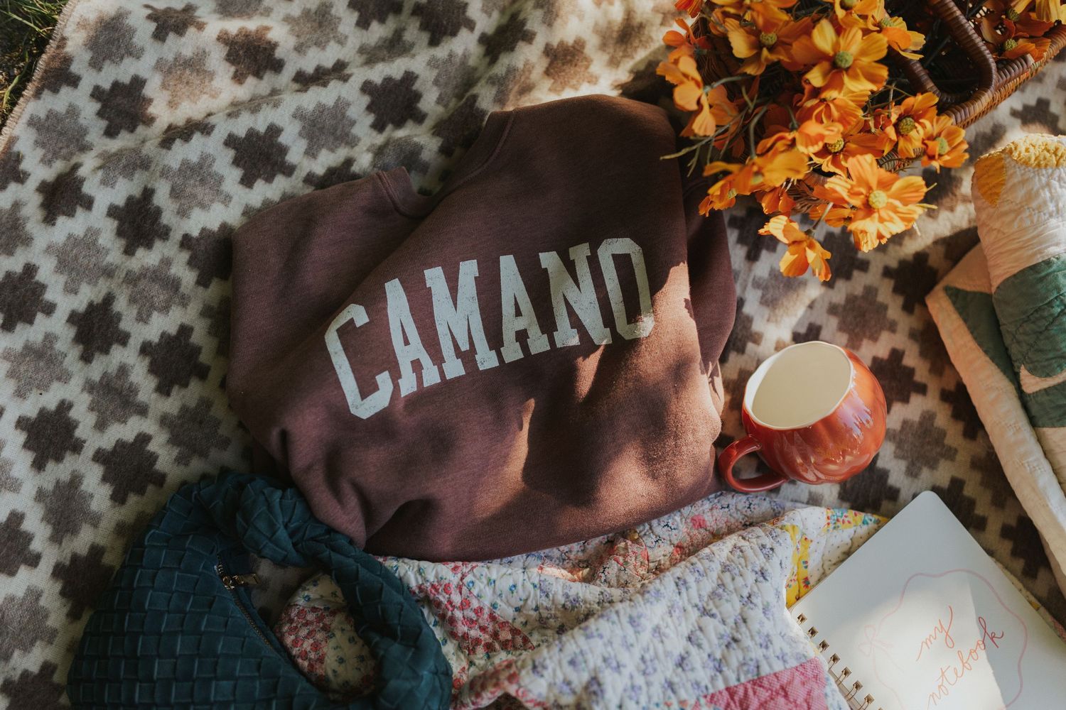 Flatlay of brown Camano sweatshirt with dried flowers on patterned blanket.