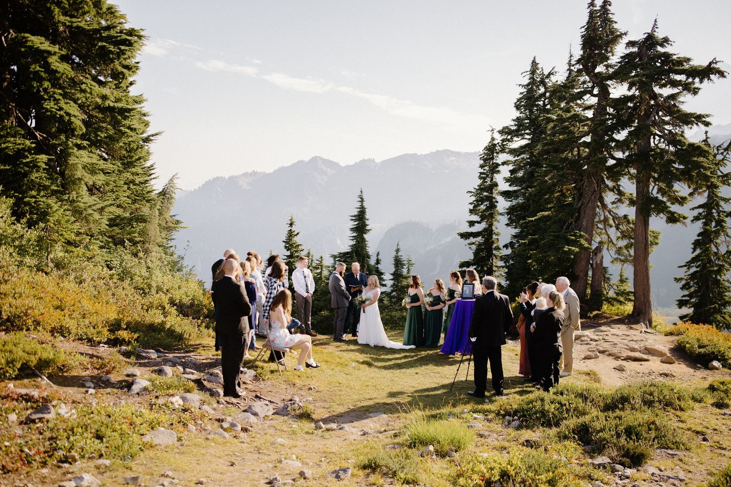 A scenic mountain wedding ceremony takes place on a hilltop surrounded by evergreen trees.