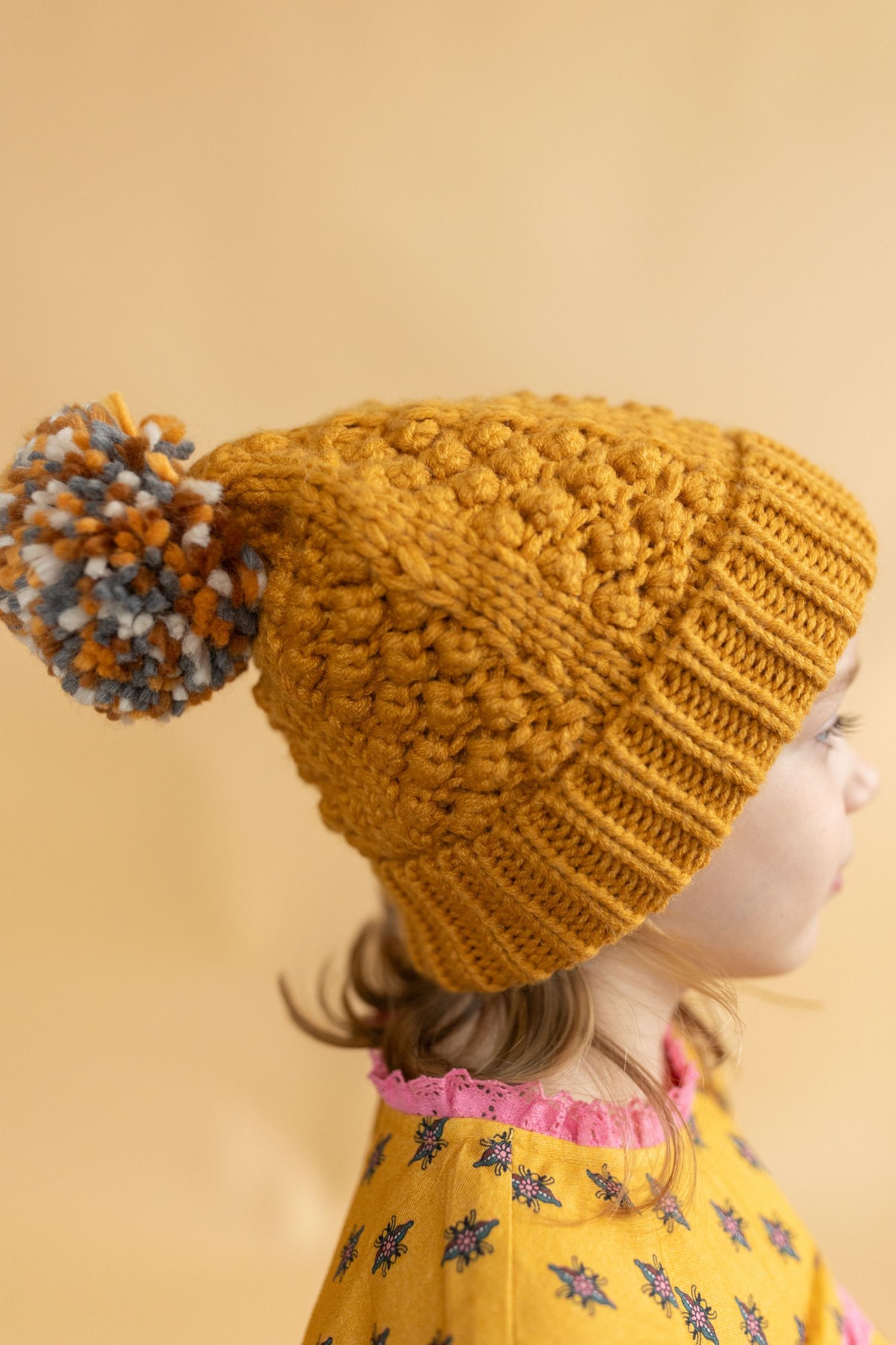 Mustard yellow knit beanie with pom pom displayed against beige background.