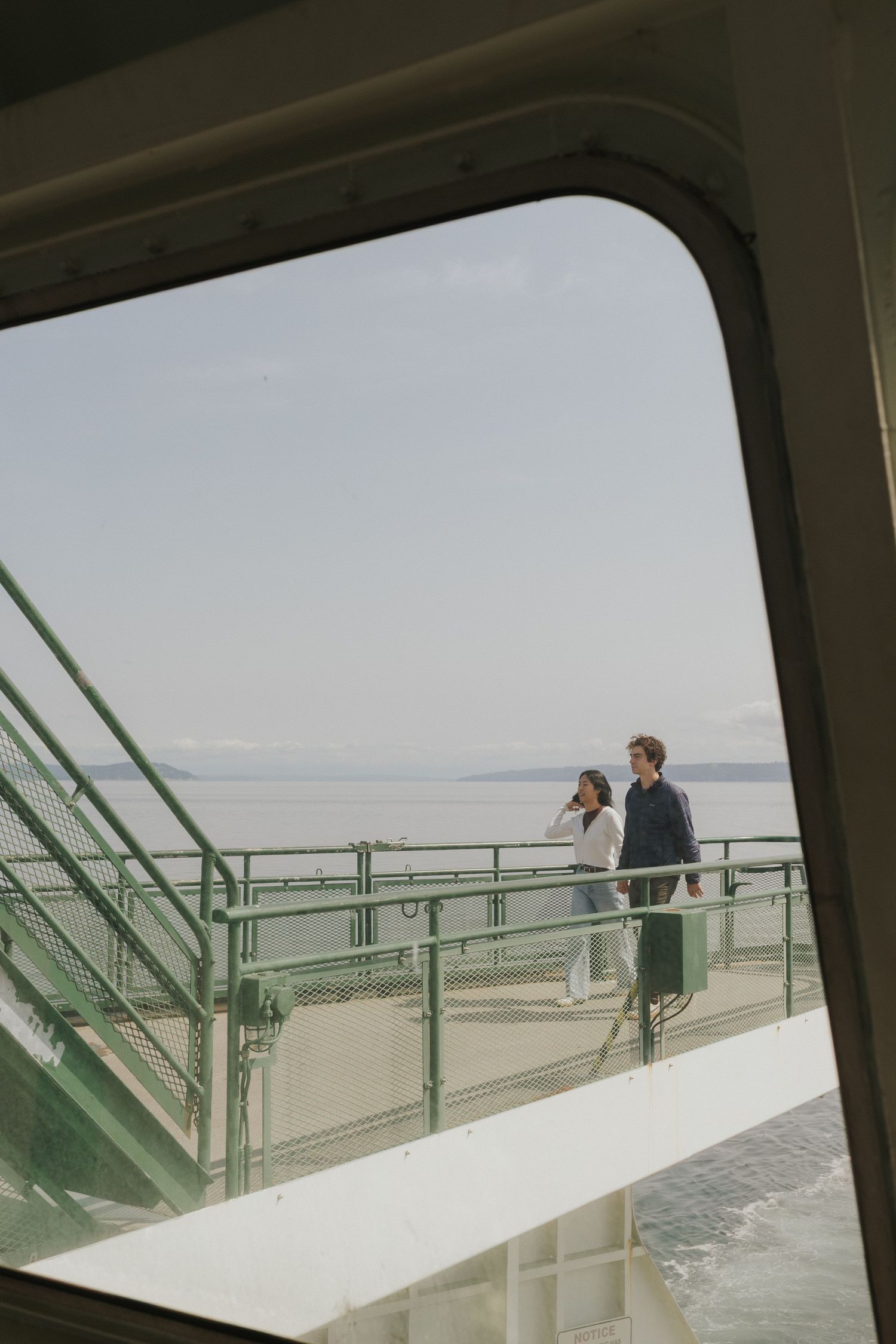 View through a ferry window of a couple standing on the observation deck against a misty horizon.