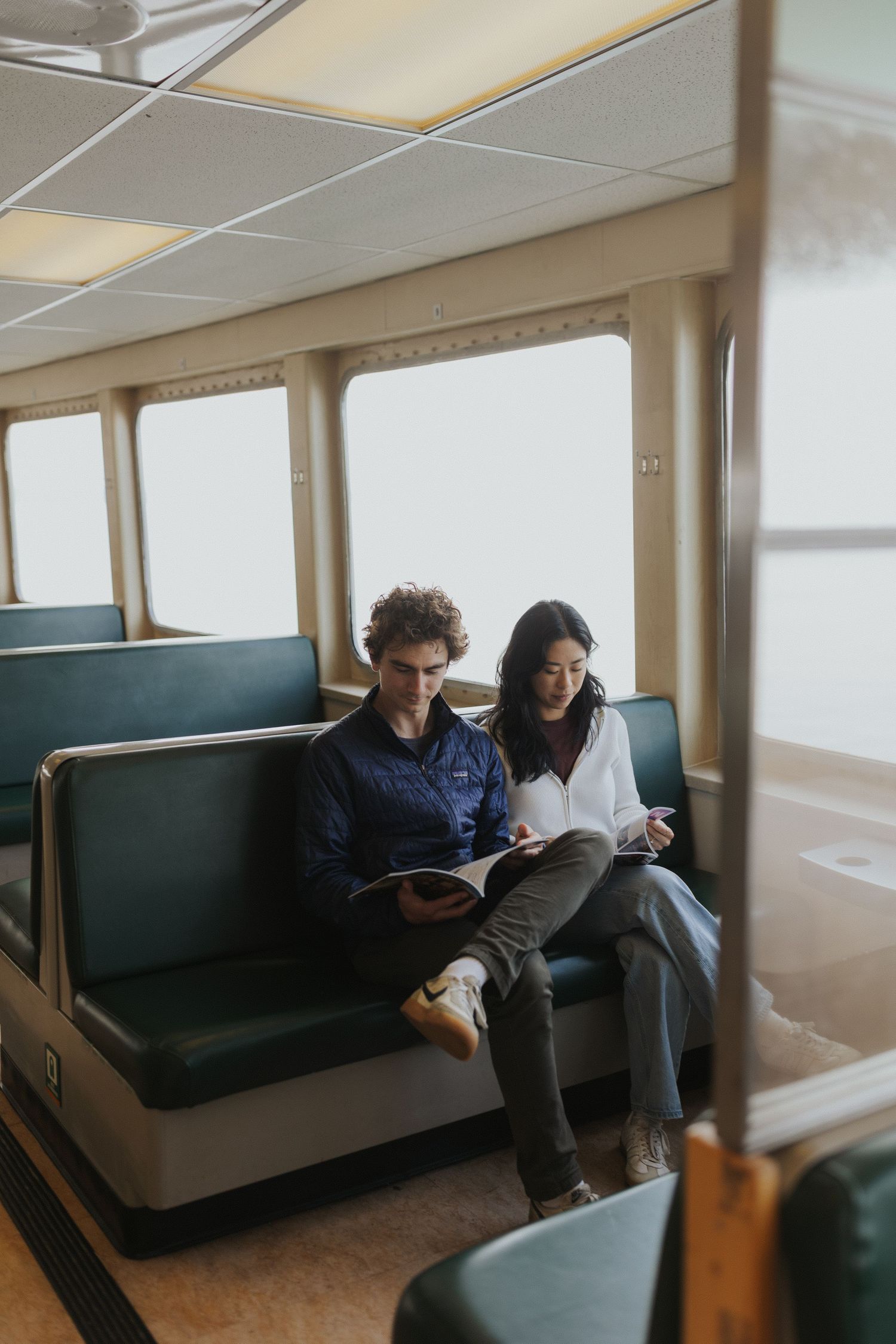 Couple relaxing on green bench seating inside ferry boat cabin.