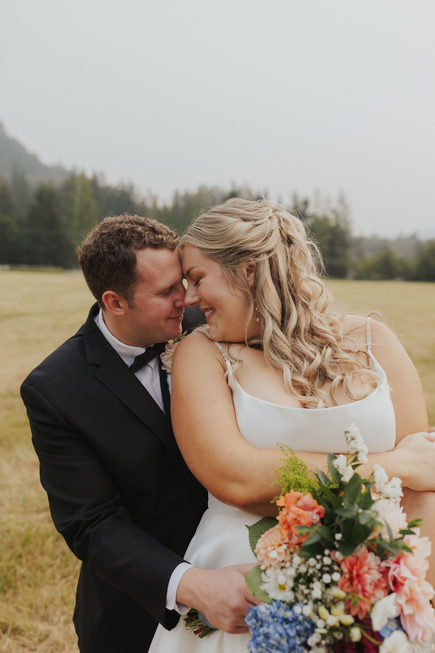 A romantic wedding couple shares a tender moment in a field while holding a vibrant bouquet of flowers.