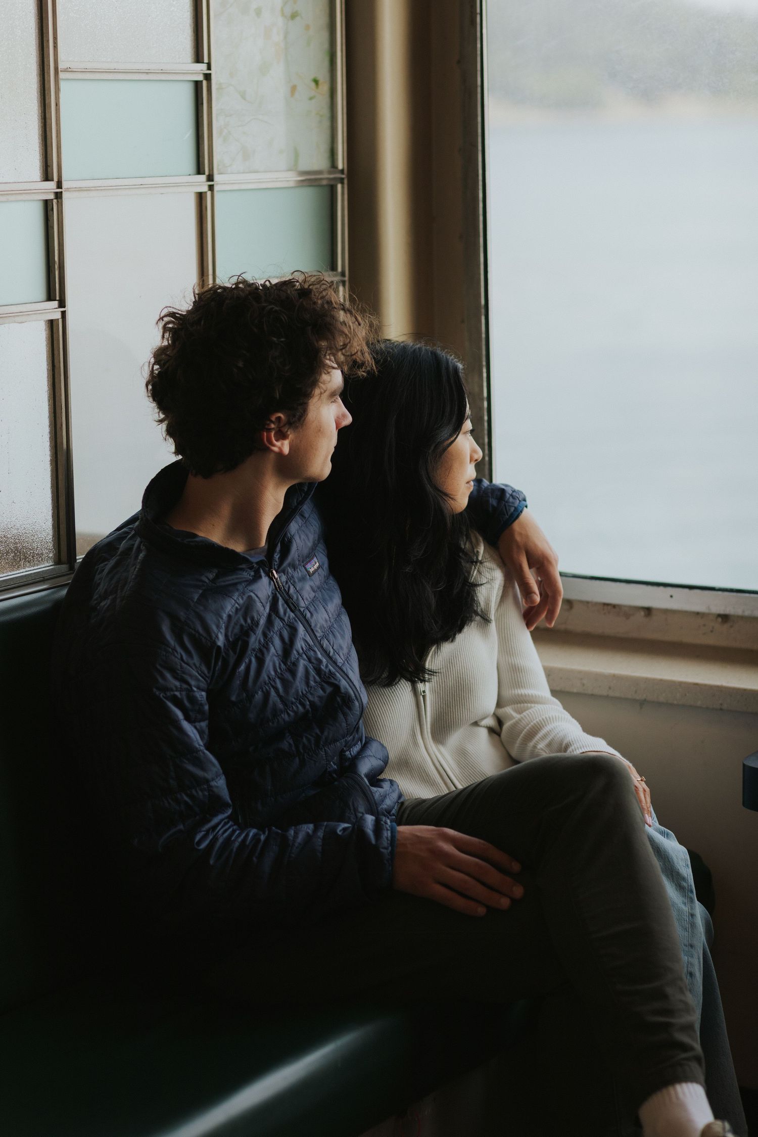 Two people sitting close together by large windows on boat, sharing quiet moment.