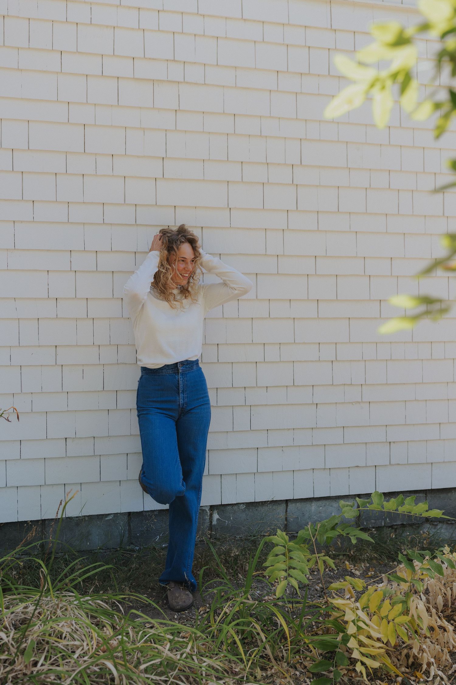 A figure leans against a white brick wall wearing blue jeans and a light colored top in a casual pose.