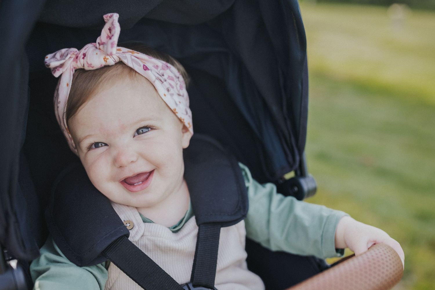 Baby in stroller smiling and wearing a floral headband outdoors on a sunny day.