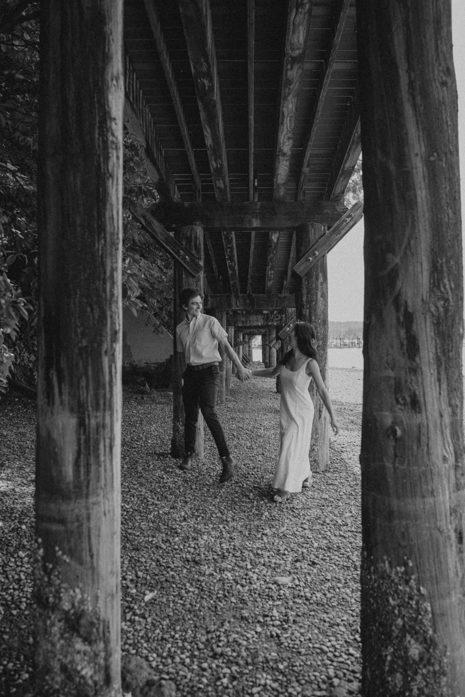 Black and white photo of couple walking under pier supports on rocky beach.