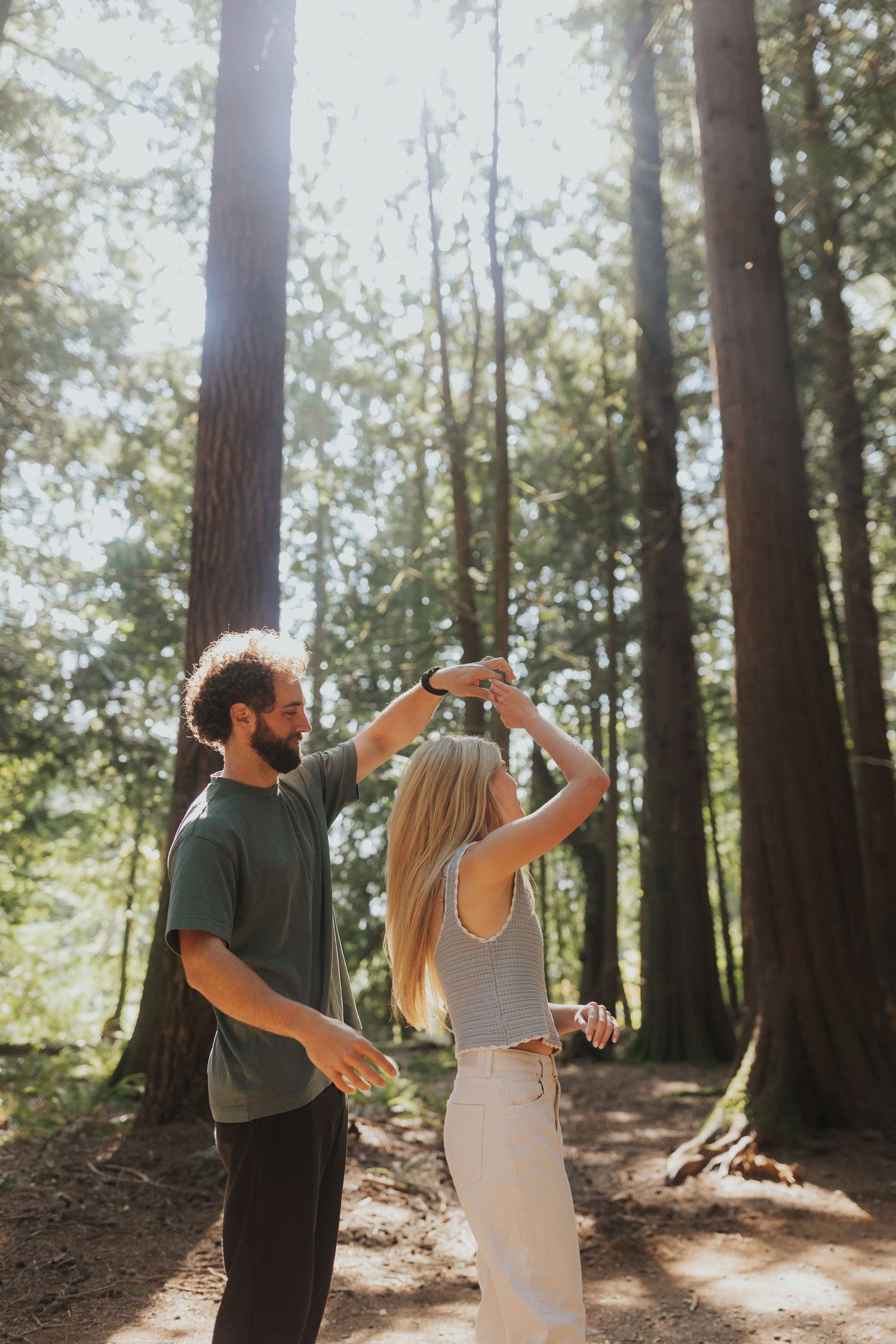 A playful couple dances in dappled sunlight filtering through towering forest trees.
