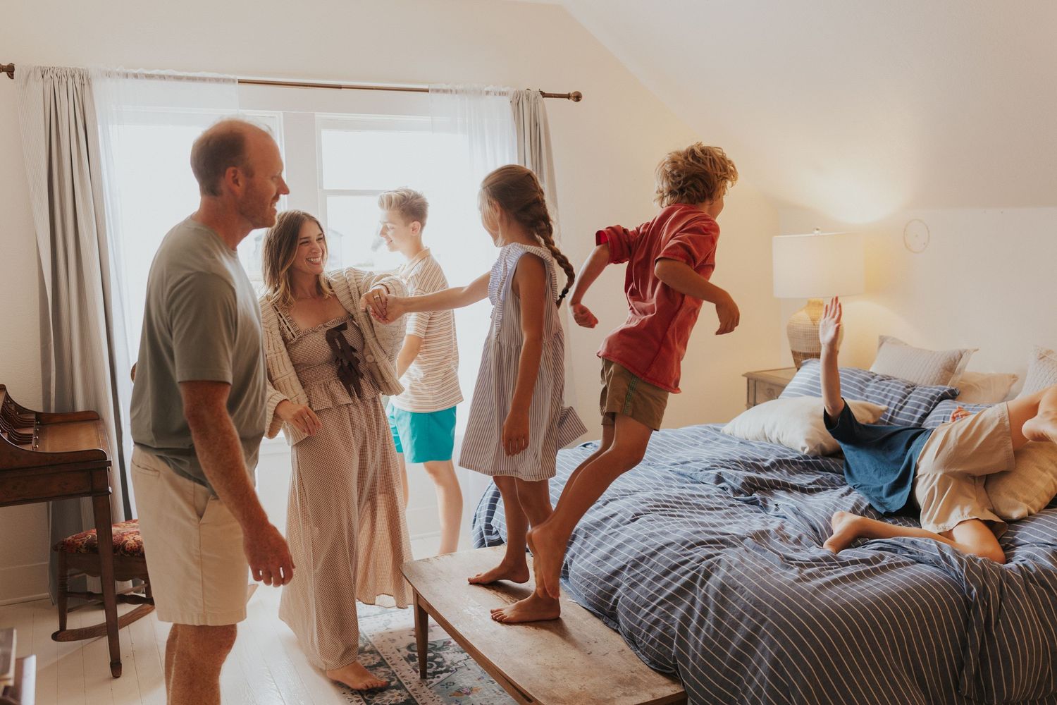 People playing and jumping on beds in a bright, sunlit bedroom.
