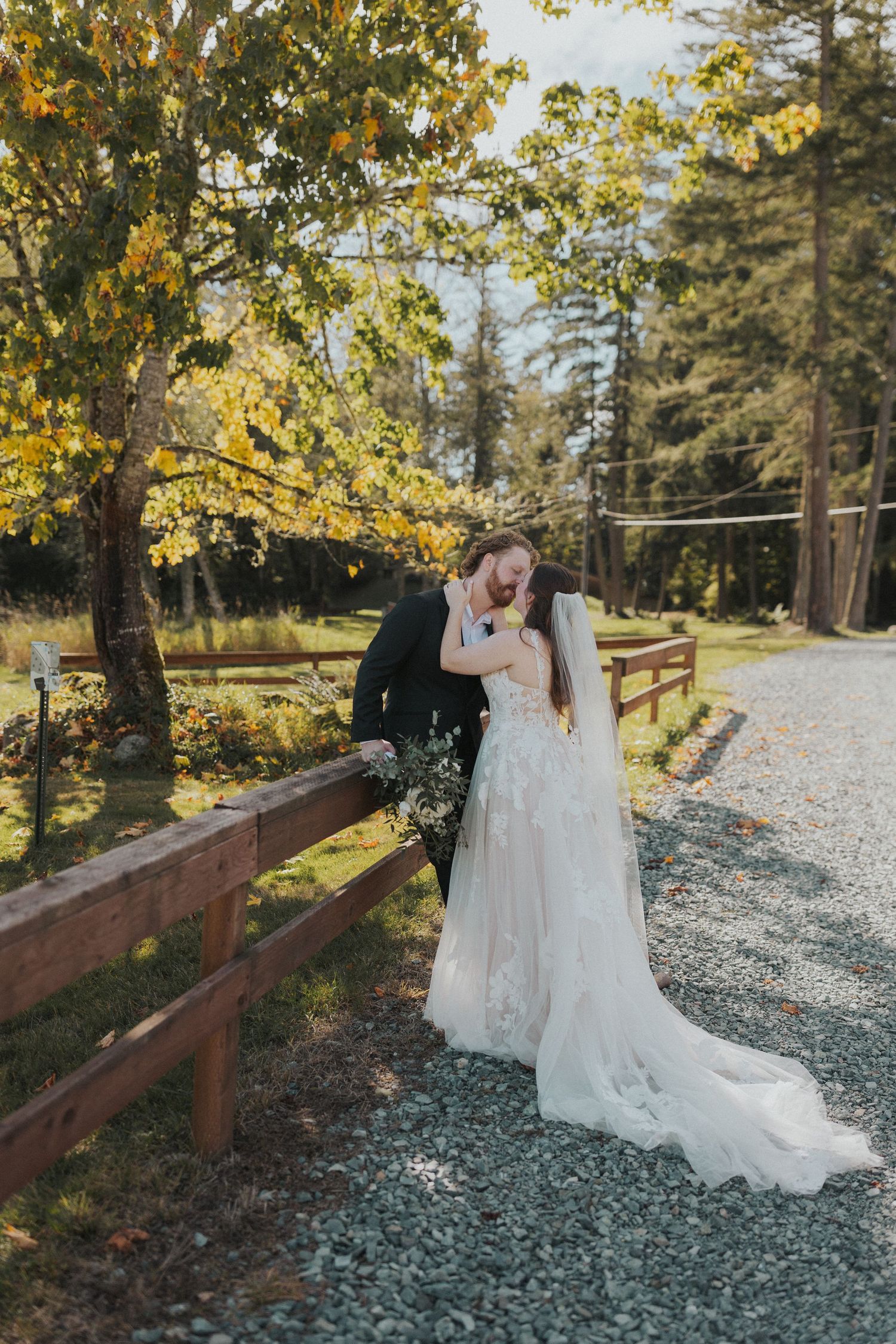Wedding couple poses along a rustic wooden fence path surrounded by fall foliage.