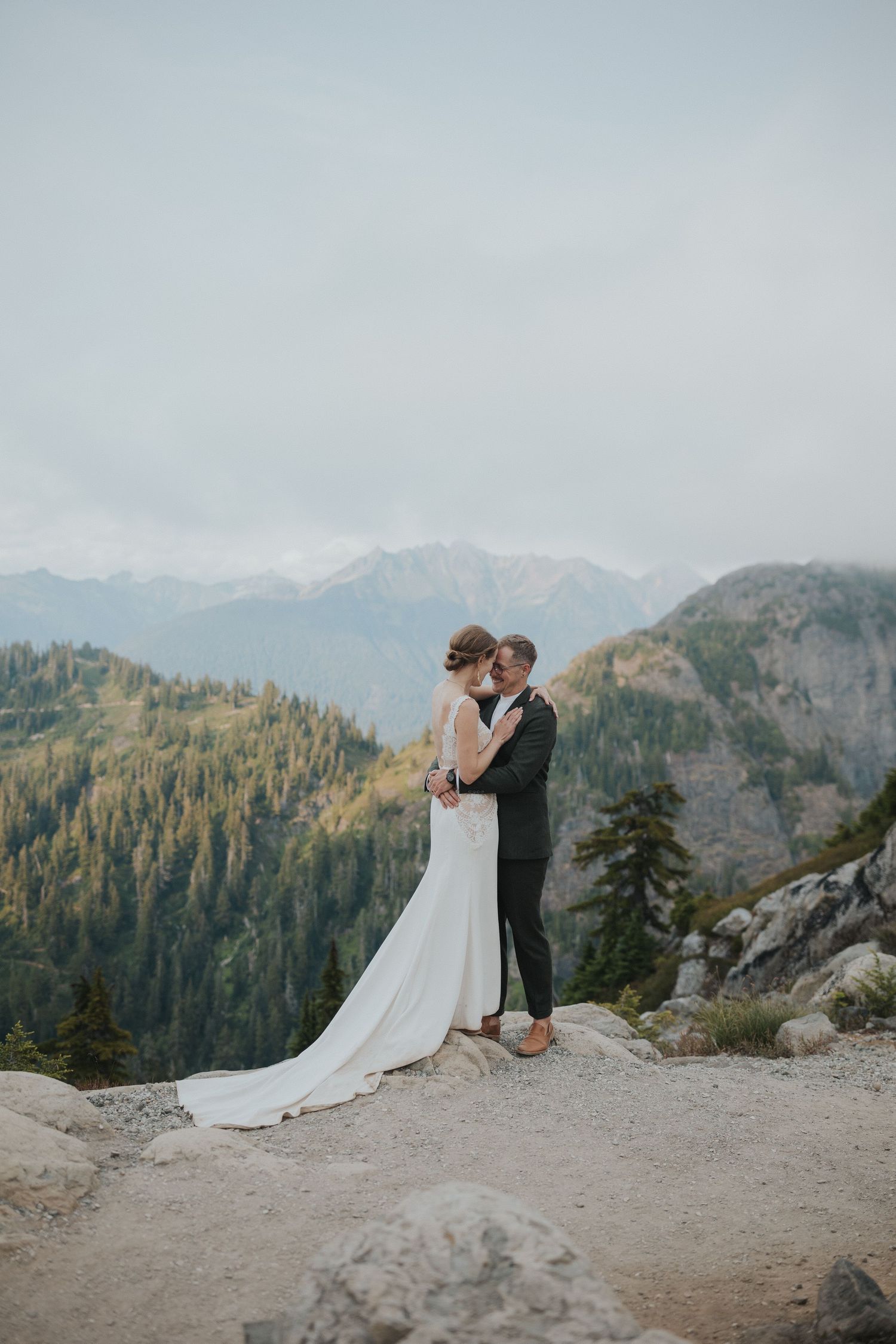 A couple shares a romantic moment on a mountain cliff overlooking evergreen forests and snow-capped peaks.