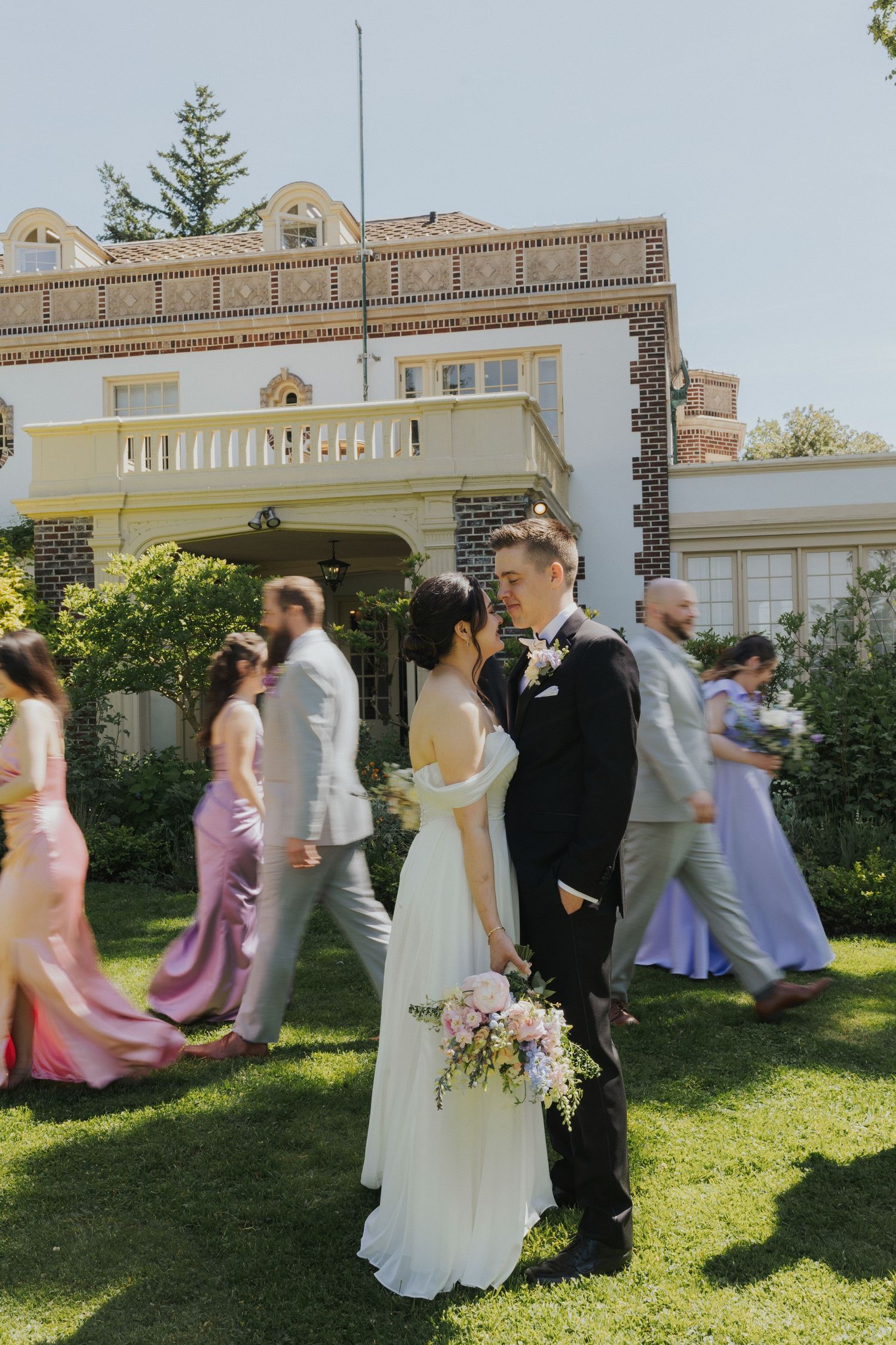 Wedding party in pastel dresses walks in line along a garden path outside a cream-colored building.