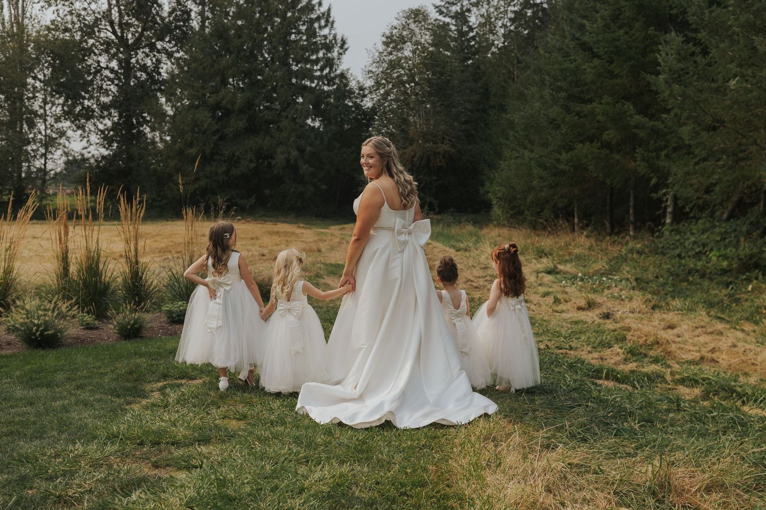 Wedding party in white dresses stands together in a sunny field with forest backdrop.