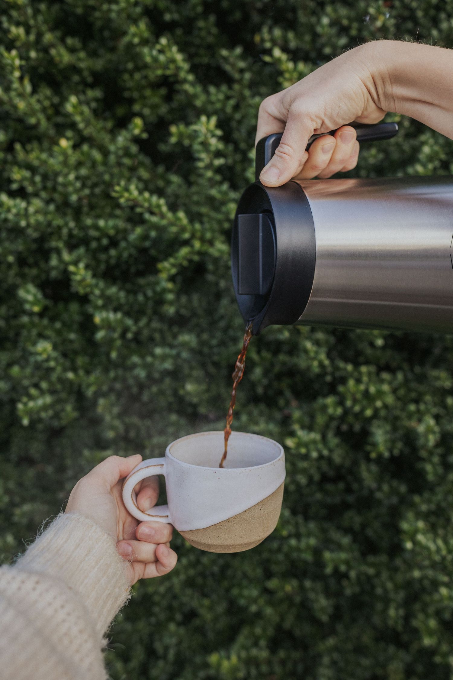 Pouring coffee from a silver thermos into a white ceramic cup against foliage.