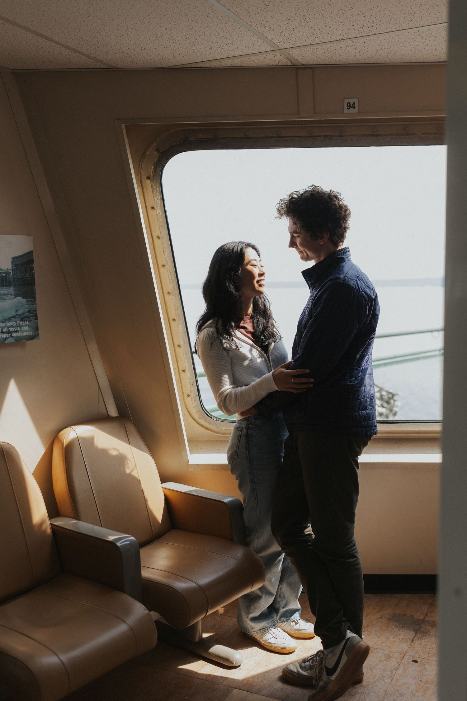 Sunlit ferry cabin interior with passengers by window seats.