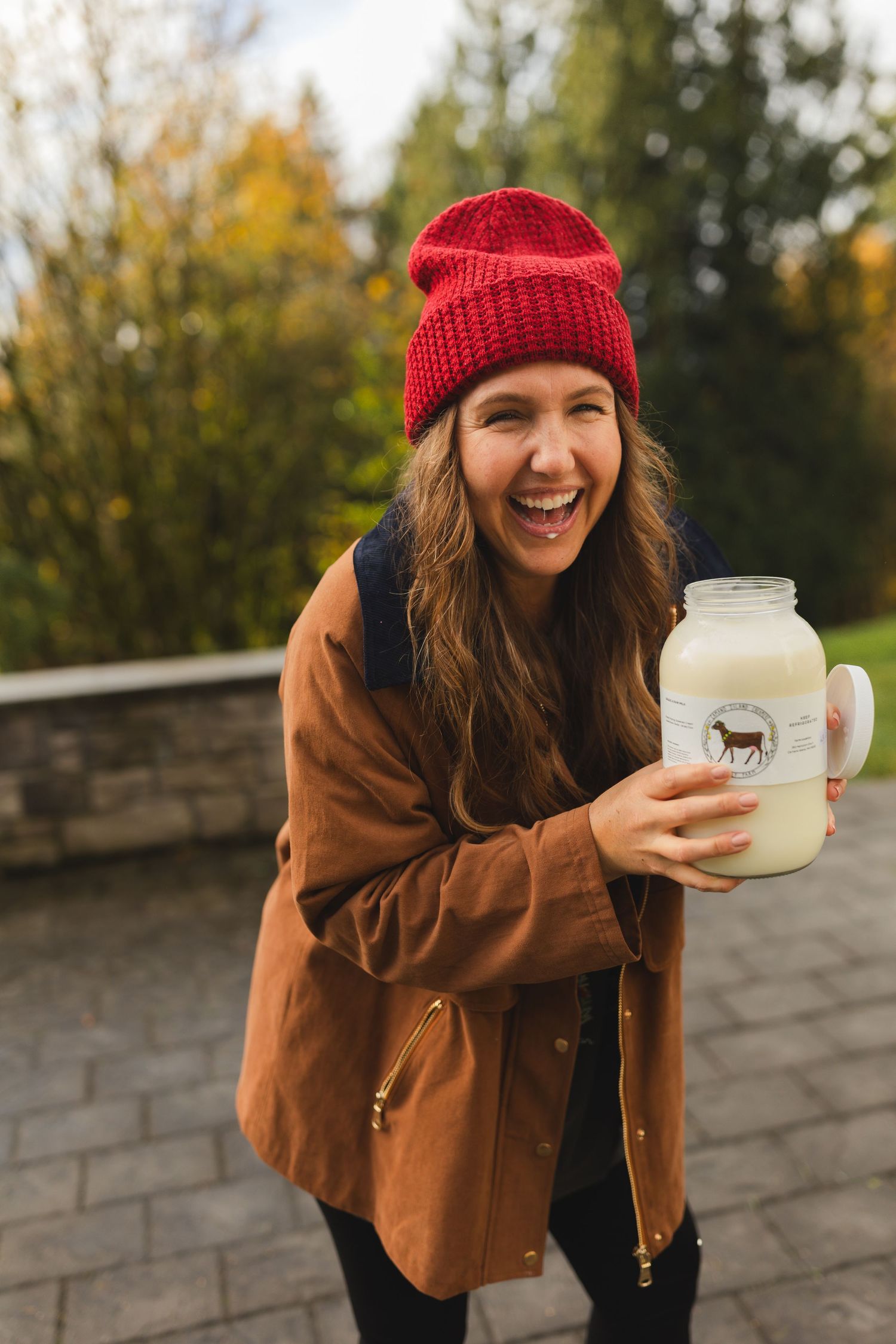 Person in brown jacket and red beanie smiling while holding a glass jar outdoors.
