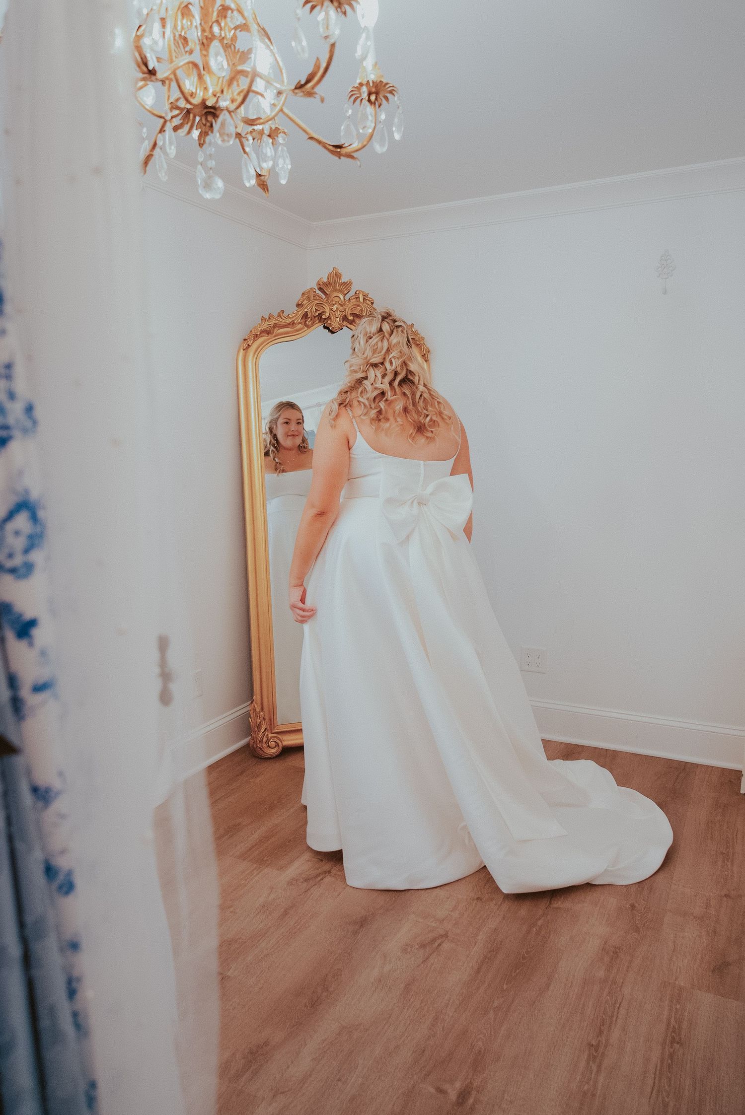 Elegant white wedding dress displayed in a room with ornate gold mirror and chandelier.