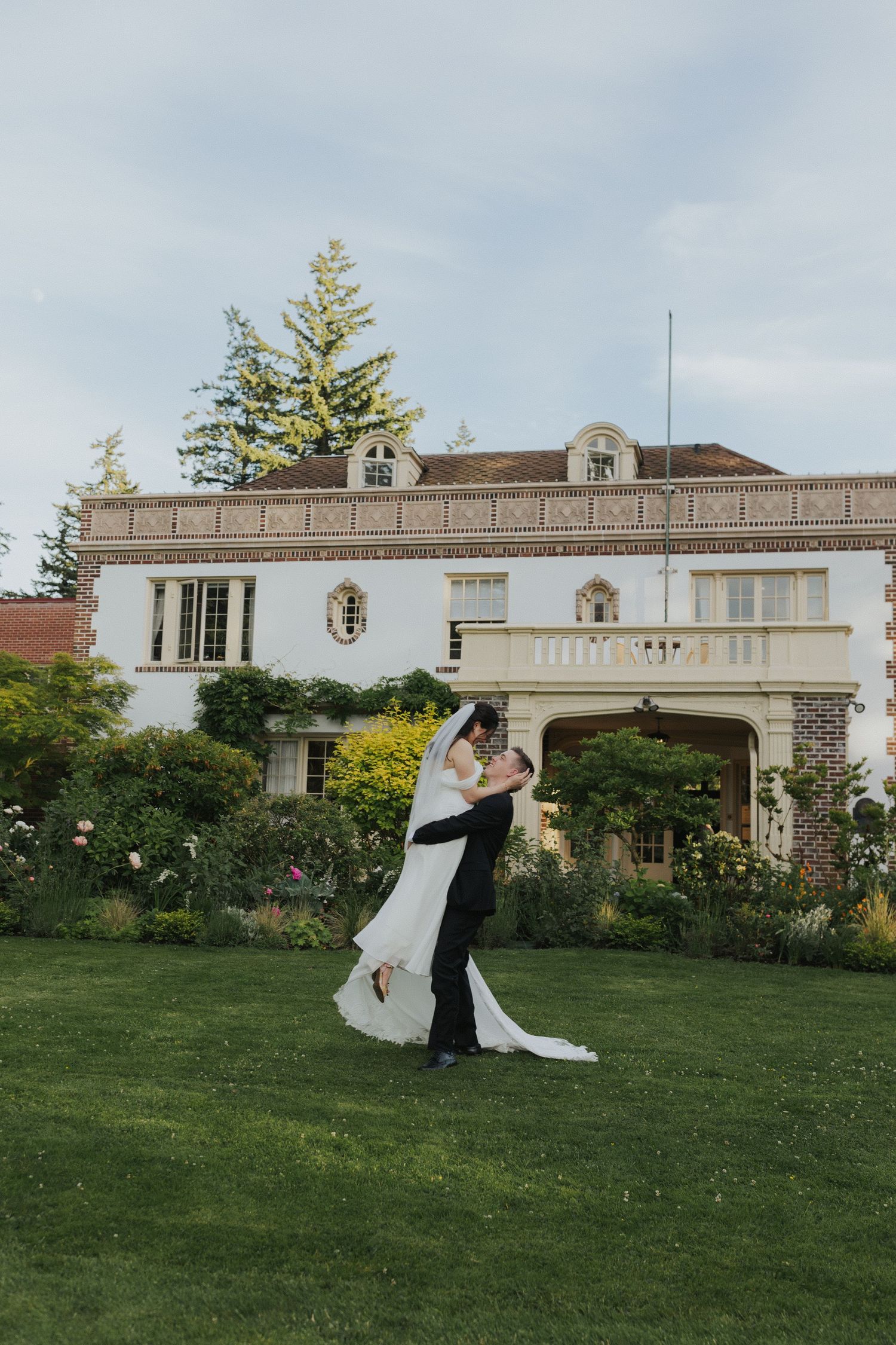 Couple in wedding attire dancing on a sprawling lawn in front of a stately white mansion.