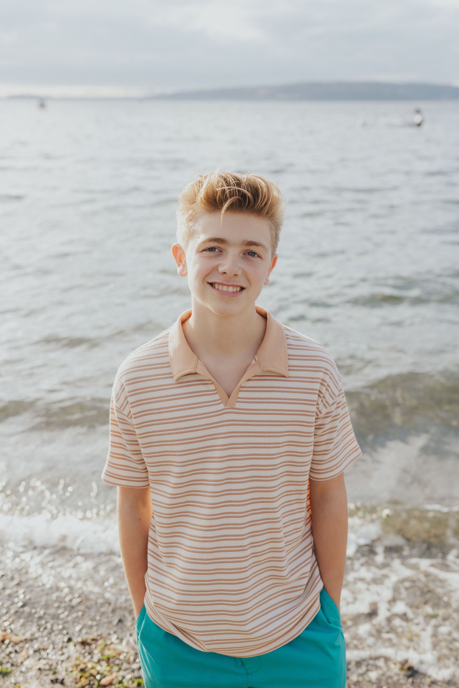 Youth in striped polo shirt smiling on beach with water in background.