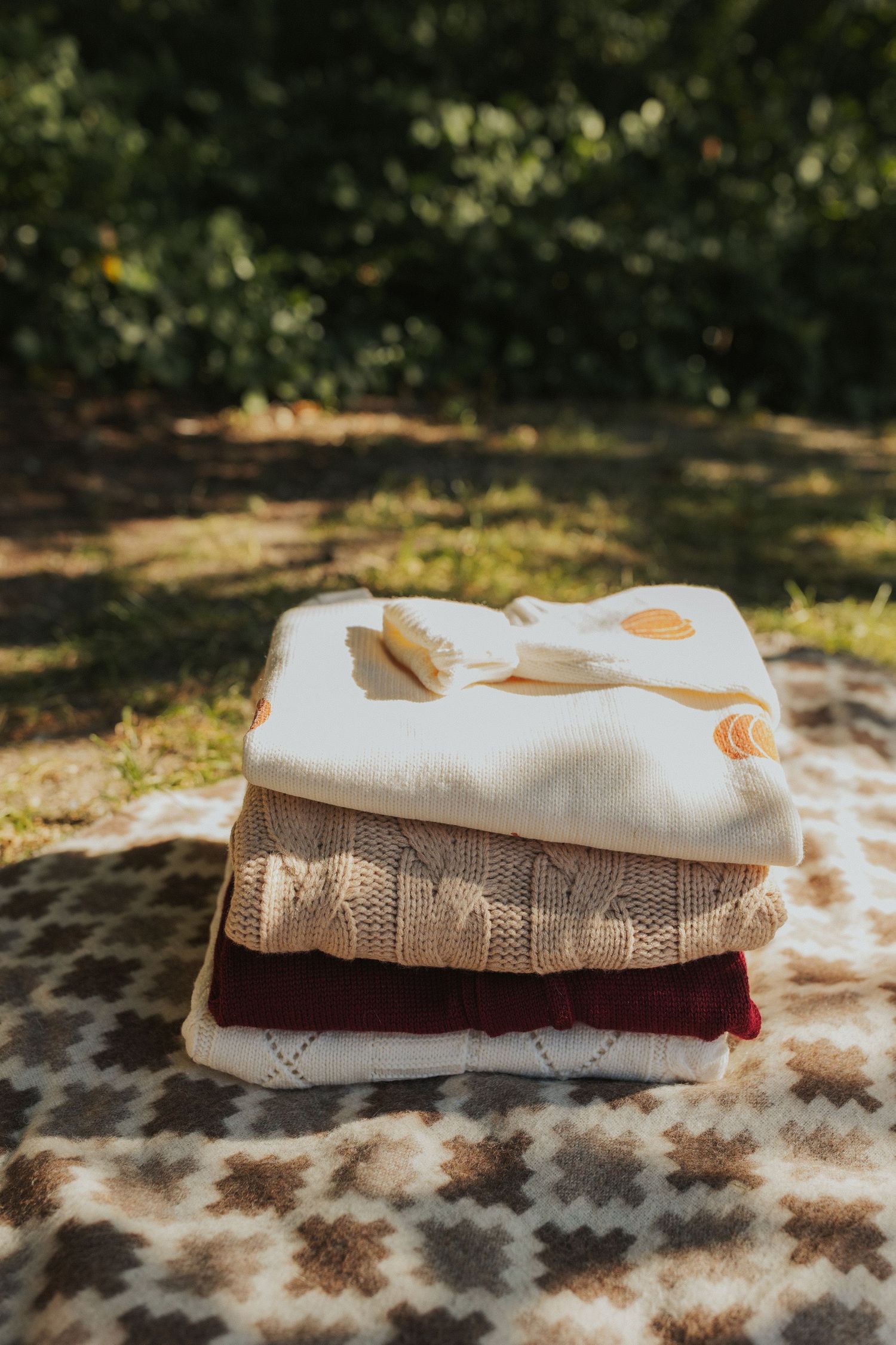 Stack of folded knit sweaters and textiles arranged on a textured surface in natural sunlight.
