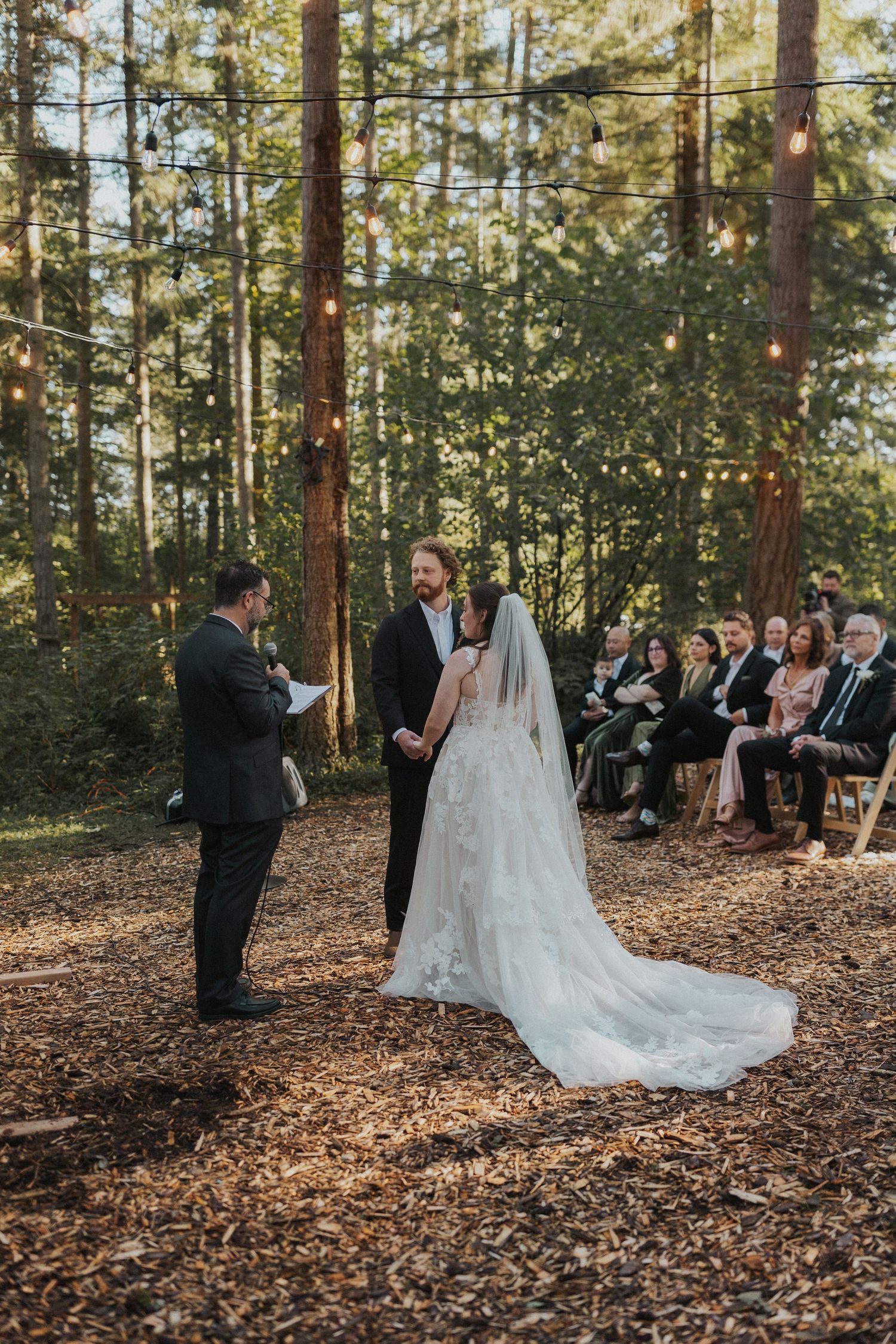 An outdoor forest wedding ceremony with guests seated among tall pine trees.