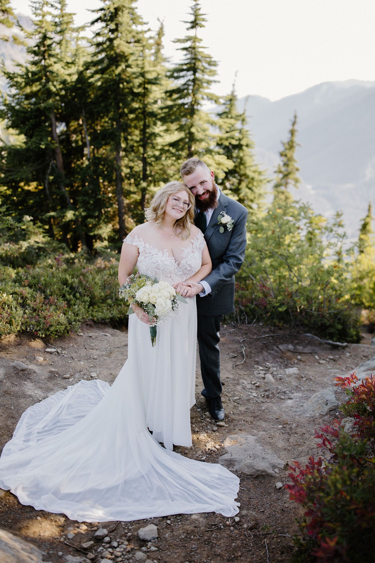 Newlyweds share an intimate moment on a mountain trail surrounded by evergreen trees.