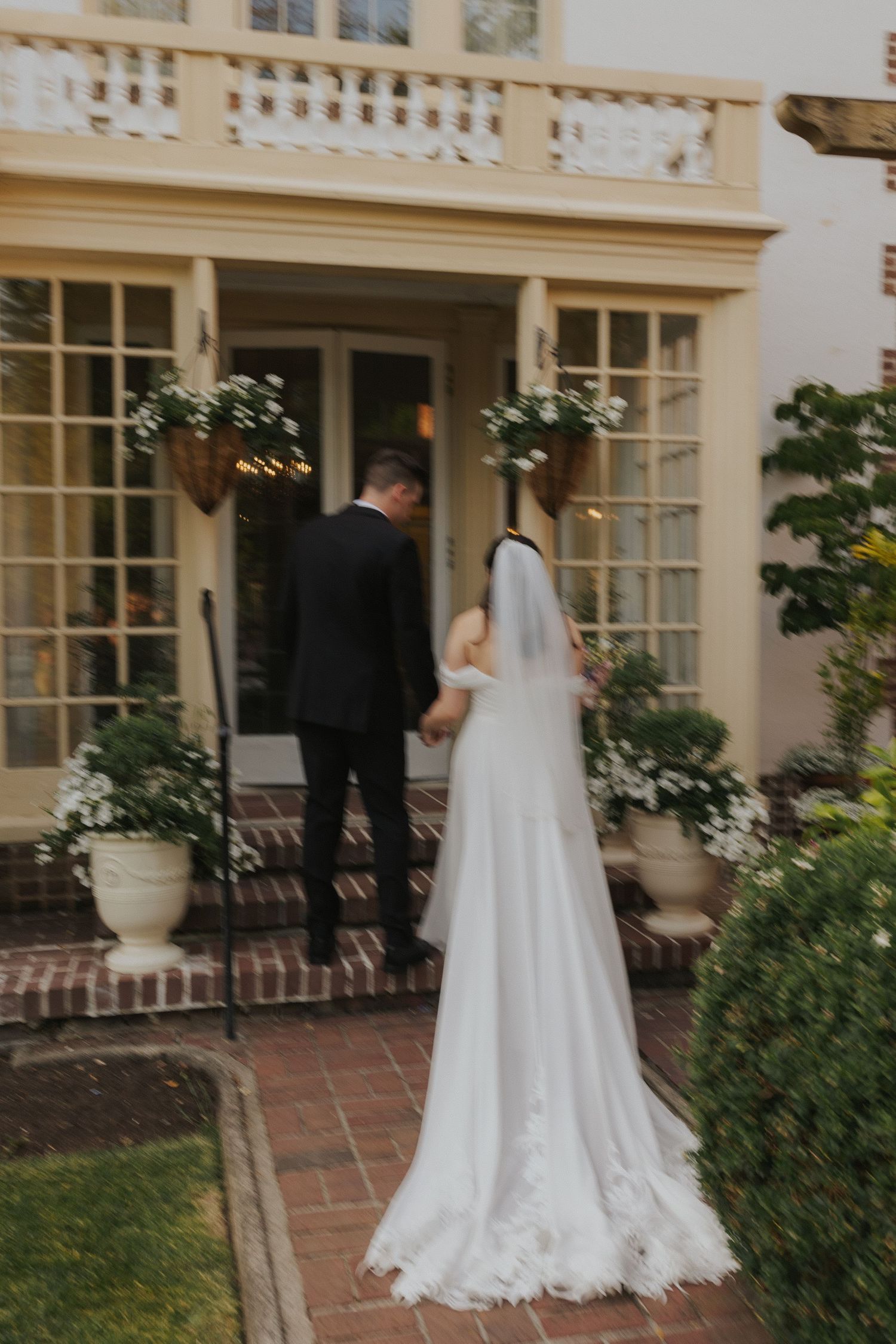 Wedding couple walking along brick pathway outside cream colored colonial style building.