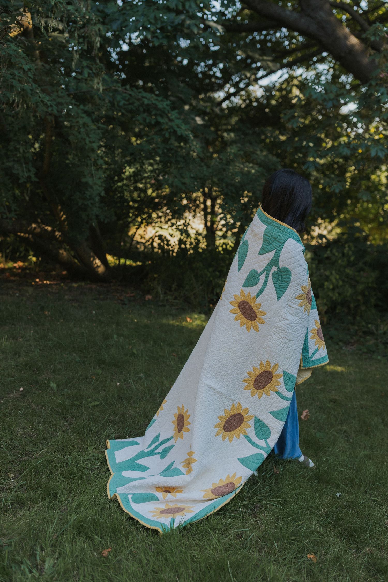 Person wrapped in white blanket with sunflower print standing in garden at sunset.