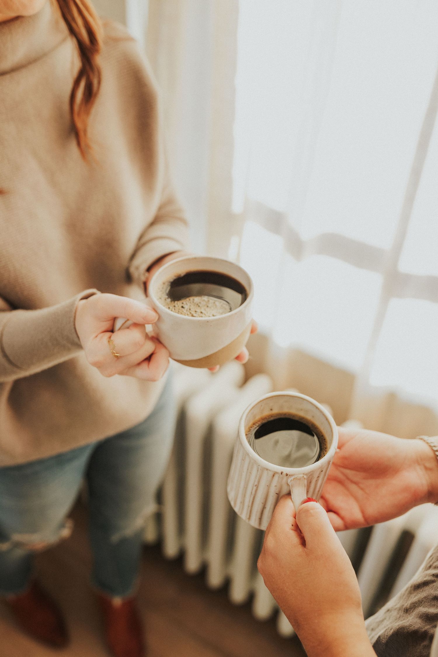 Hands holding coffee cups in warm sunlight through window.