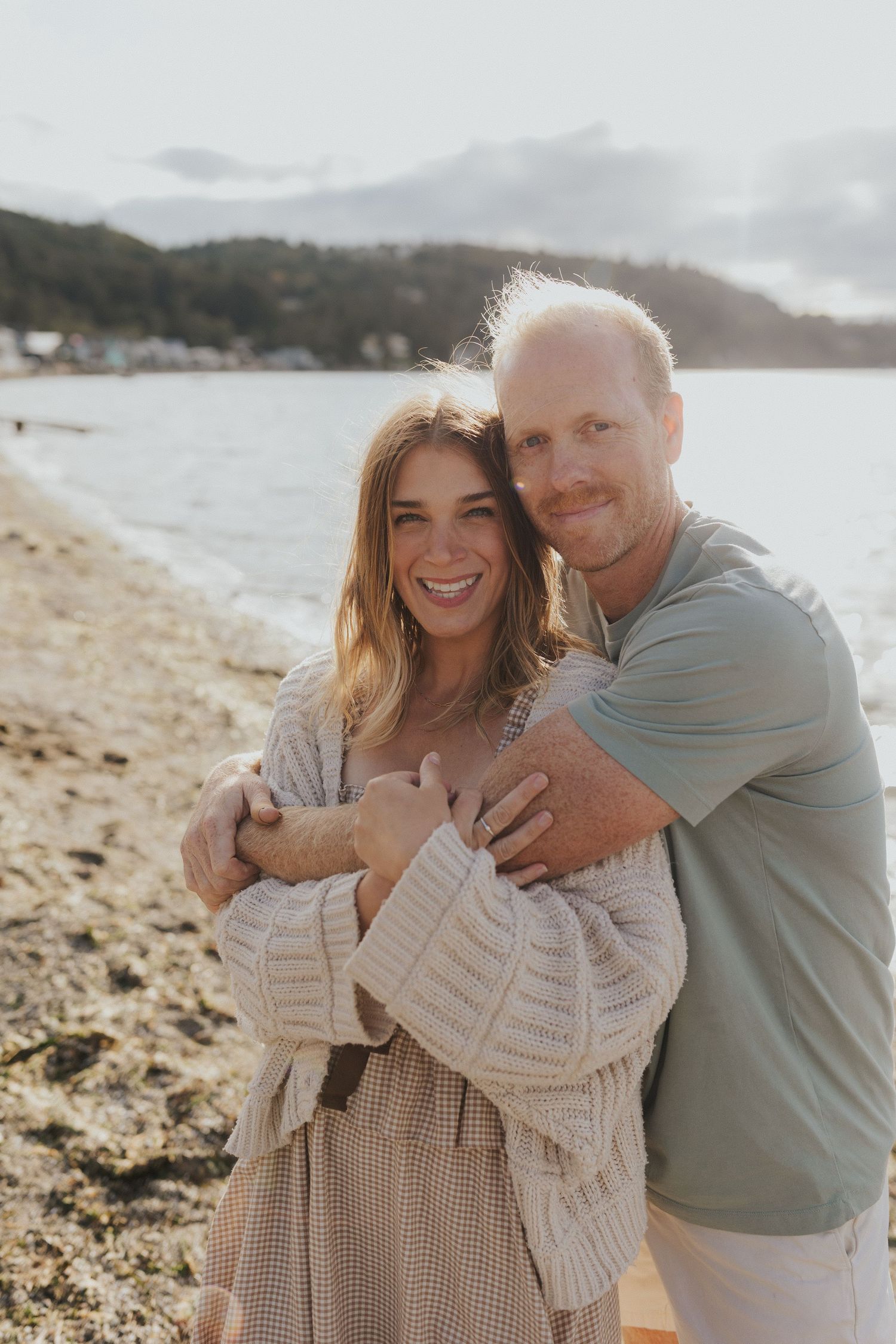 Couple embracing on a scenic beach with mountains and water in the background.